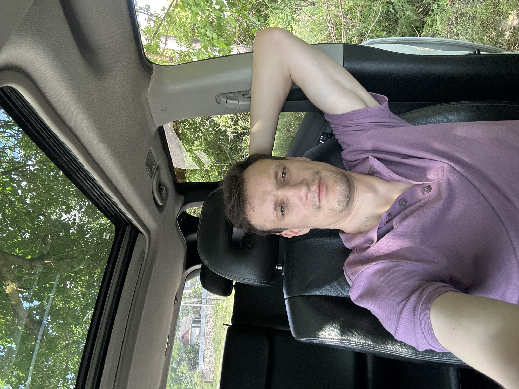 A man with short brown hair and stubble is seated in the driver's seat of a car, looking at the camera. He is wearing a purple polo shirt and has his right arm resting behind his head. The car has a sunroof, which is open, revealing trees and a blue sky. The interior of the car is visible, including black leather seats. Outside the car, there are green trees and foliage, and a glimpse of a house and a stone wall. The scene appears to be in a rural or suburban area, consistent with the village of Makiv, Ukraine.