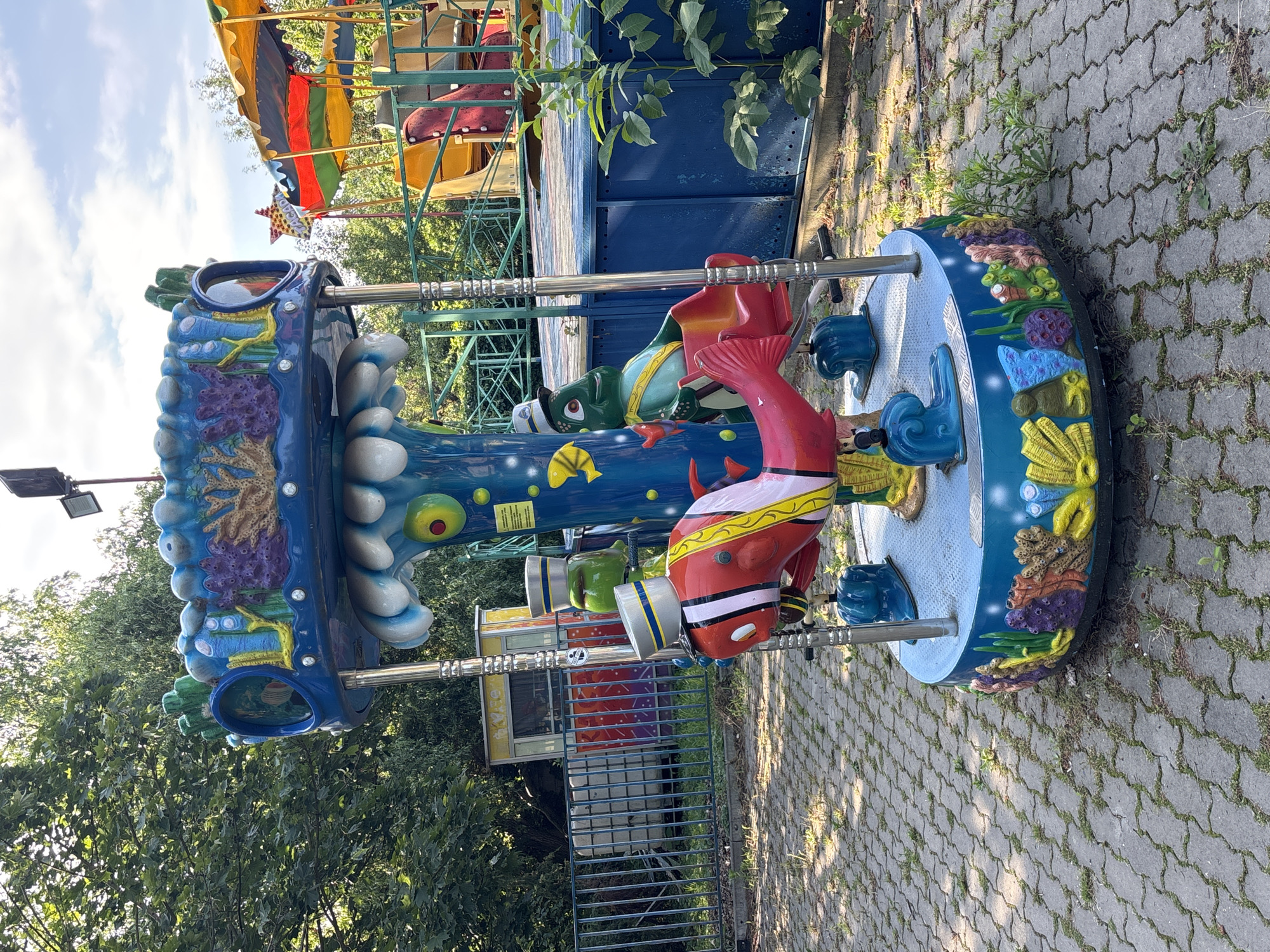 An outdoor, eye-level, medium shot captures a children's carousel in an amusement park or fairground. The carousel has a nautical theme, with a central pole painted blue and decorated with marine elements like coral, seaweed, and stylized waves. Attached to the pole are three seats shaped like cartoonish sea creatures: a red fish with white stripes and a sailor's hat, a green turtle-like creature, and another green creature, all wearing sailor hats. The seats are red and have a somewhat worn appearance. The base of the carousel is also adorned with colorful reef decorations.
The carousel is situated on a paved area made of gray interlocking pavers, with some patches of grass and weeds growing in between. In the background, a large blue structure with a yellow and red striped canopy can be seen, possibly another ride or concession stand. Behind this, a chain-link fence encloses the area, and further back, dense green trees line the perimeter. A small, weathered yellow booth with a window and colorful graphics is visible behind the fence. The sky is partly cloudy with some blue sky visible. The lighting suggests it is daytime, likely late morning or early afternoon, given the shadows cast. There are no people visible in the image.