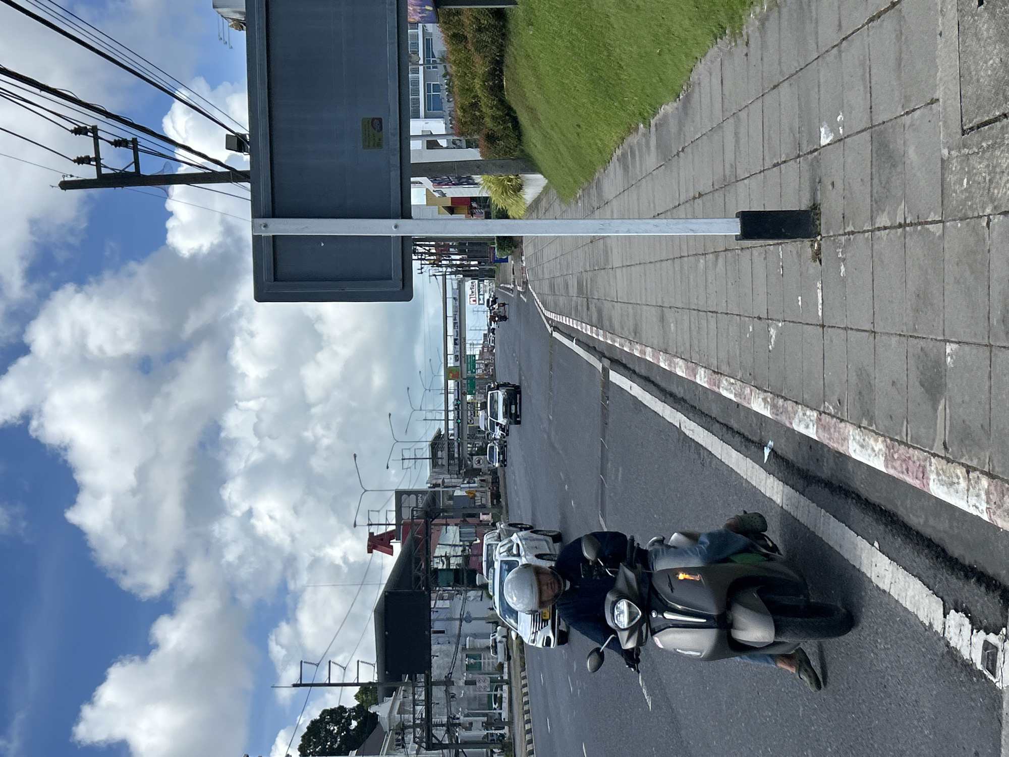 An overhead shot shows a man riding a dark grey scooter on a street in Wichit, Thailand. He is wearing a white helmet, a dark blue jacket, and blue jeans. The scooter has a headlight and a yellow turn signal. The man is on the left side of the frame, moving towards the background. The scooter is in the foreground, occupying the lower left quadrant of the image.
The street is asphalt with white and red painted lines. A paved sidewalk with gray square tiles runs along the right side of the street, bordered by a grassy embankment and a large, dark grey rectangular sign. The sky above is blue with scattered white clouds. In the background, there are various buildings, including a white building with a red "A" sign, power lines, streetlights, and other vehicles on the road. The overall scene depicts a busy street in a Thai city on a cloudy day.