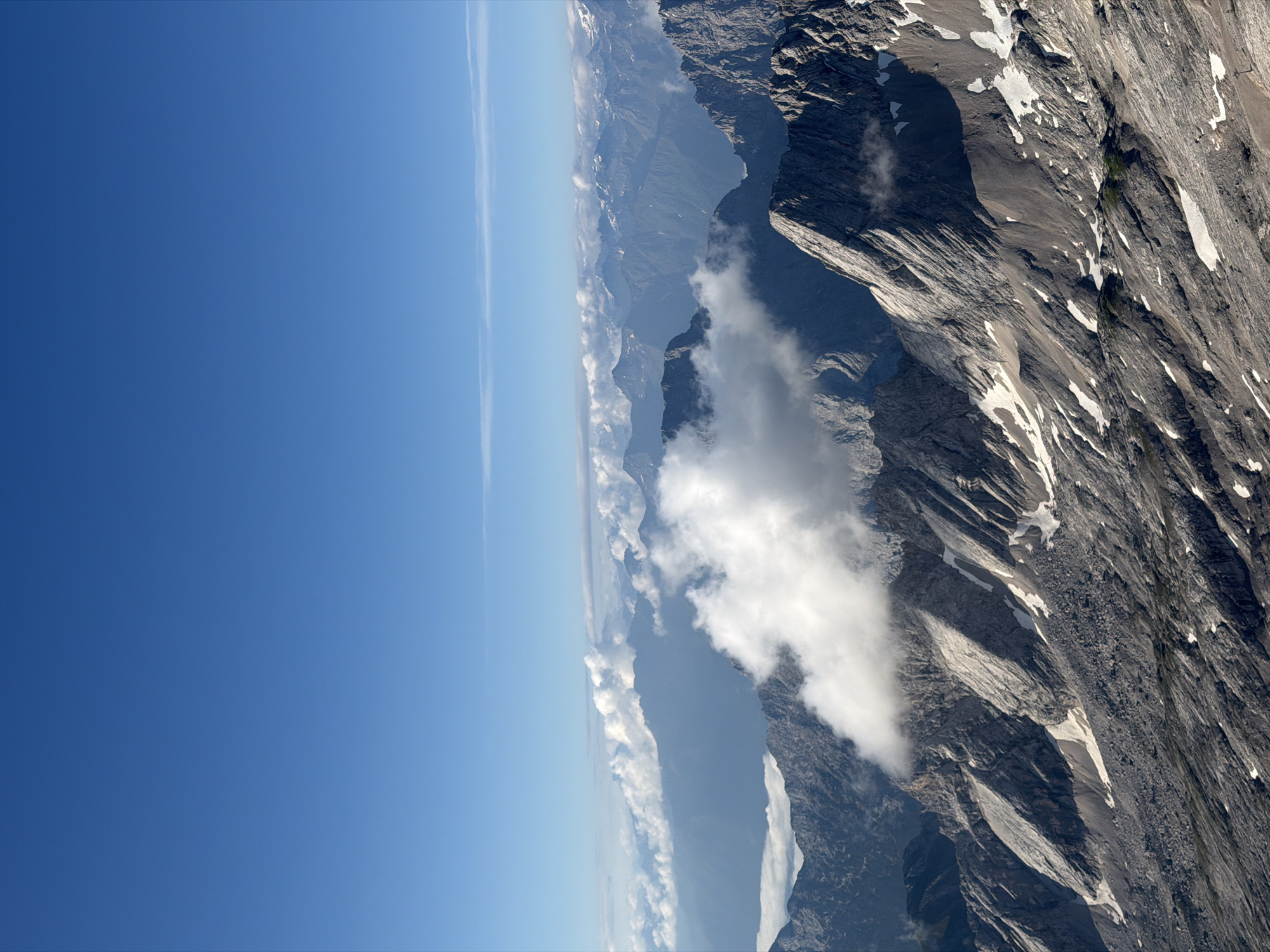 This is an aerial, wide-angle vertical photograph of a mountain range, likely taken from a high vantage point such as a drone or aircraft, above the town of Grainau, Germany. The sky is a clear, vibrant blue, with a thin, horizontal band of clouds in the distance. The foreground is dominated by rugged, rocky mountain slopes, with patches of snow clinging to the shaded areas and crevices. The terrain is uneven and appears to be composed of scree and rock formations. In the mid-ground, a large, fluffy white cloud partially obscures a section of the mountain peaks, creating a dramatic contrast with the dark grey rock. Further in the distance, a series of progressively fainter mountain ranges can be seen, fading into a hazy blue silhouette against the sky. The overall impression is one of immense scale and natural, wild beauty. There are no people or man-made structures clearly visible in the image.