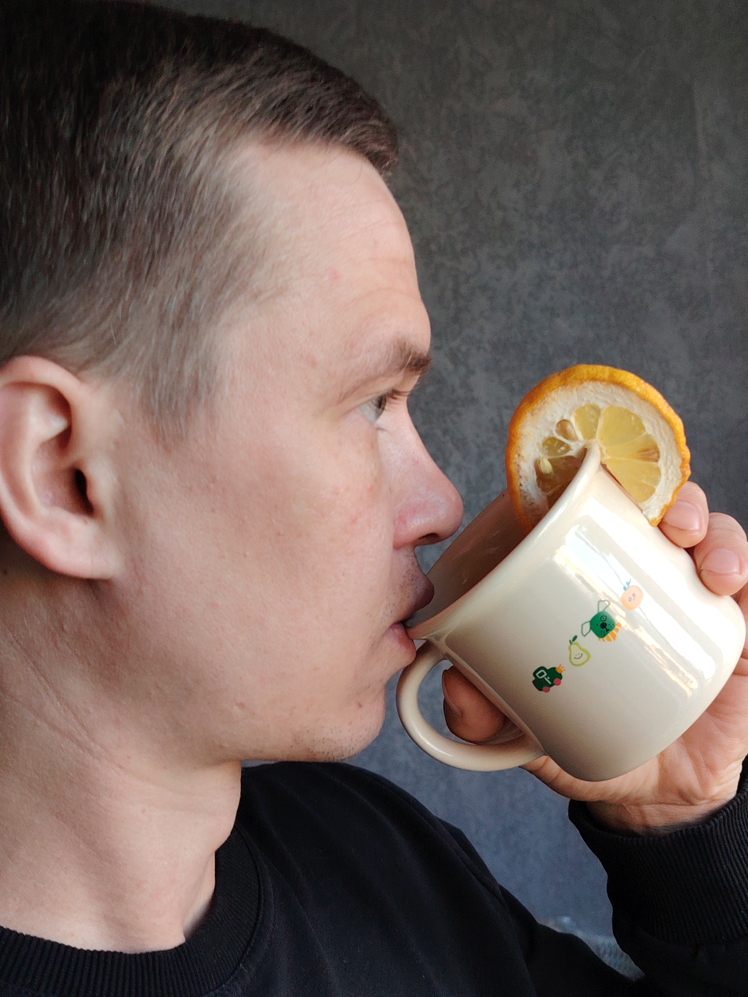 A man is drinking from a mug with a lemon slice on the rim. The mug has cartoon images of a pear, a green dog, and a toy car. The man is wearing a black sweatshirt. The background is a mottled gray. No information is available about the location of this image within the city of Chaikovsky, Russia.