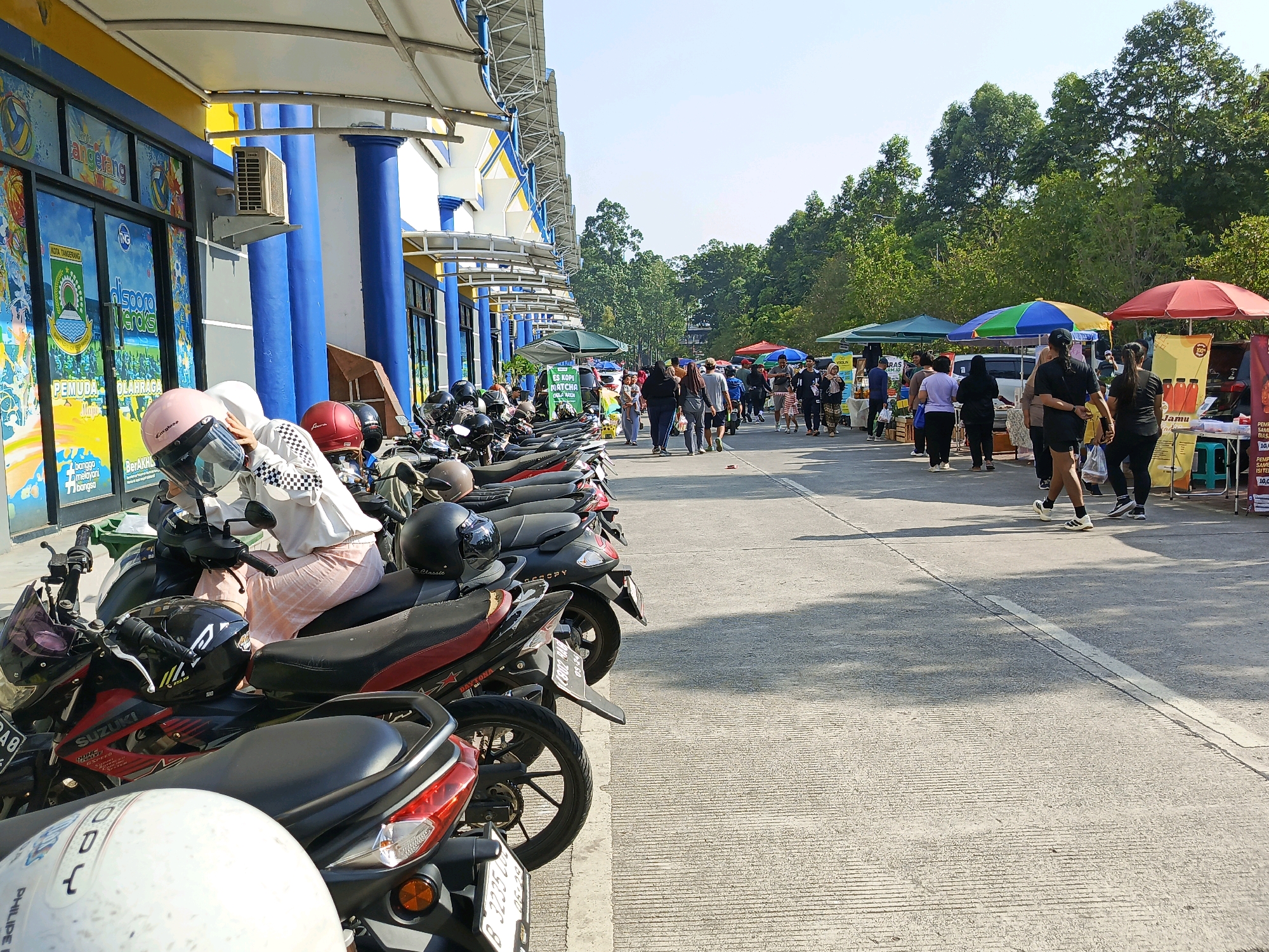 The image depicts an outdoor scene in Tangerang, Indonesia, likely a marketplace or a public event area adjacent to a building. In the foreground, a row of parked motorcycles, some with helmets on them, are visible. A person is seated on one of the motorcycles, wearing a light-colored top and a pink helmet. The building in the background has a blue and yellow facade with large windows displaying graphics. Further into the scene, a paved pathway is lined with stalls or vendors selling various goods, indicated by colorful umbrellas and tables. Several people are walking along the pathway, browsing the stalls or engaging in social interaction. The environment appears to be sunny with a clear sky, and trees are visible in the distance, suggesting a warm climate.