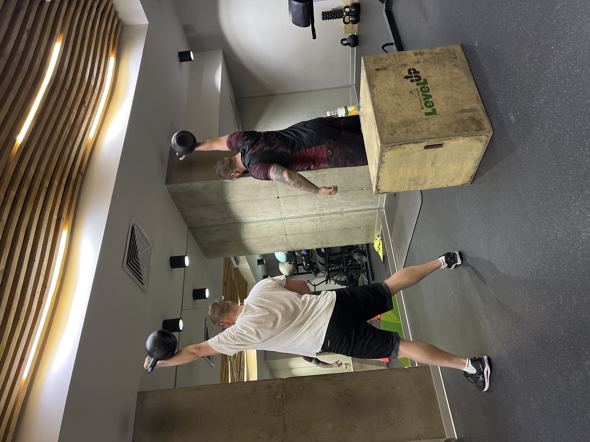 In a gym in Krasnoyarsk, Russia, two men are performing kettlebell exercises. The man on the left, facing away from the camera, wears a white t-shirt and black shorts. He is lifting a black kettlebell above his head with his right arm. The man on the right, also facing away, wears a dark red and black t-shirt and dark shorts. He is lifting a black kettlebell above his head with his right arm. Both men are standing on a dark gray rubber floor. In the foreground, a wooden plyometric box with the "LevelUP" logo is visible. The gym environment includes concrete walls, mirrors, gym equipment in the background, and a wooden slatted ceiling with integrated lighting.