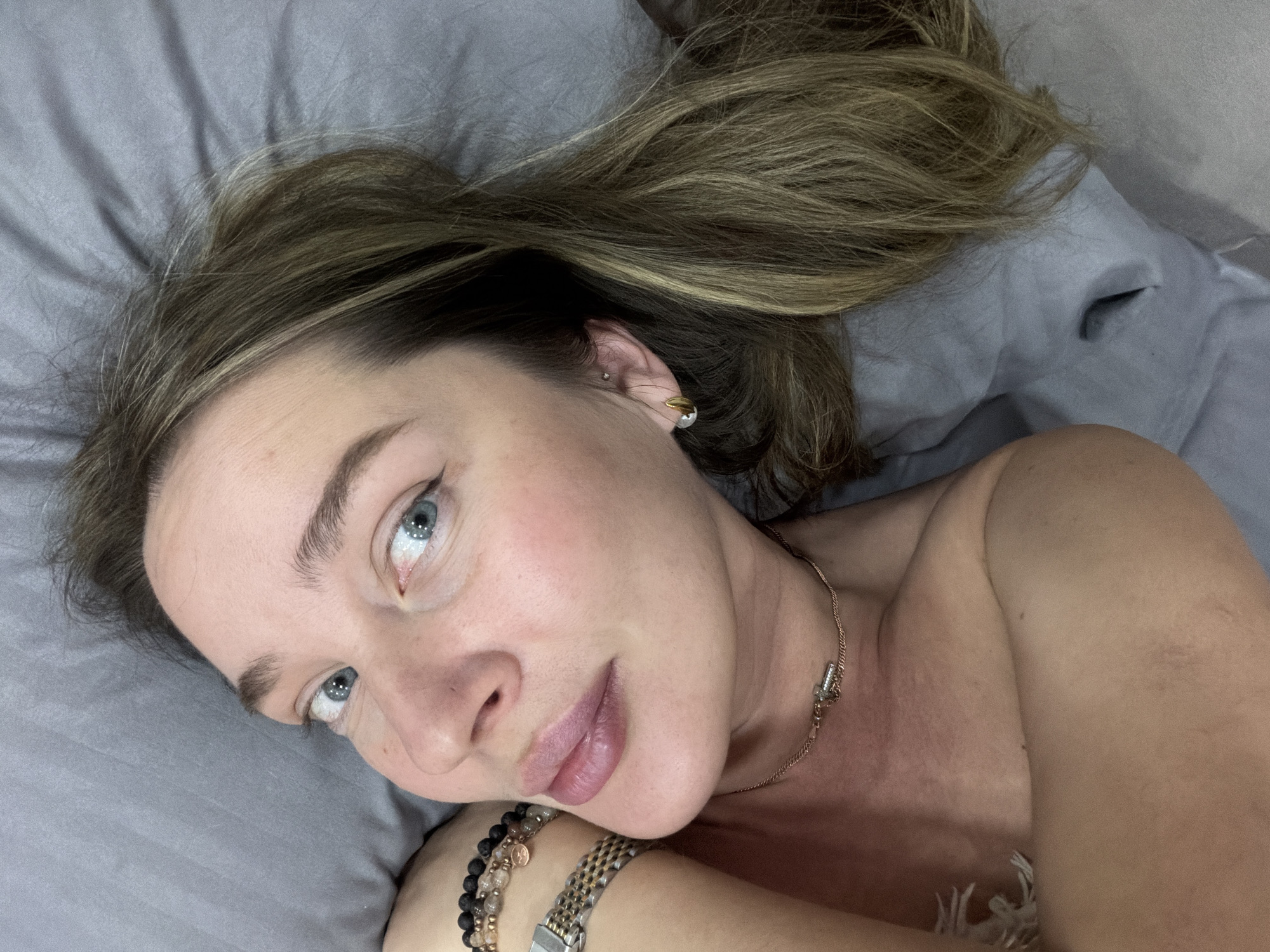 A woman with fair skin, light brown hair with blonde highlights, and blue eyes lies on a gray bed. She is wearing a gold necklace and bracelets. The location is not evident, so its existence in the city of Chaikovsky, Russia, cannot be verified.