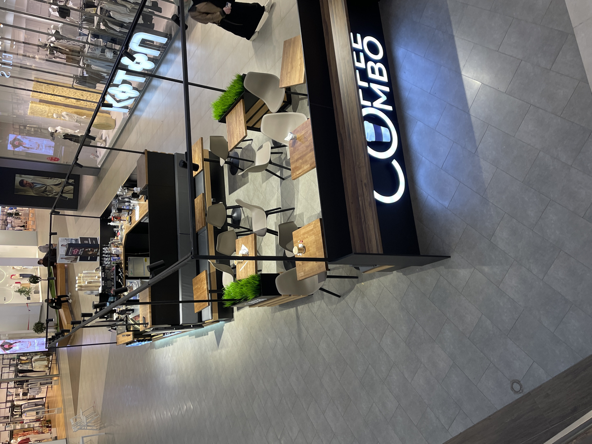 The image shows an indoor shopping mall environment in Shcherbinka, Russia. The scene features a "Coffee Combo" kiosk. The kiosk has a black and brown color scheme, and its name is prominently displayed vertically in illuminated letters. Adjacent to the kiosk, there are several tables and chairs arranged for customers. The flooring is gray tiles. Various retail clothing stores are visible in the background behind glass displays and mannequins. A person is seen walking away from the kiosk.