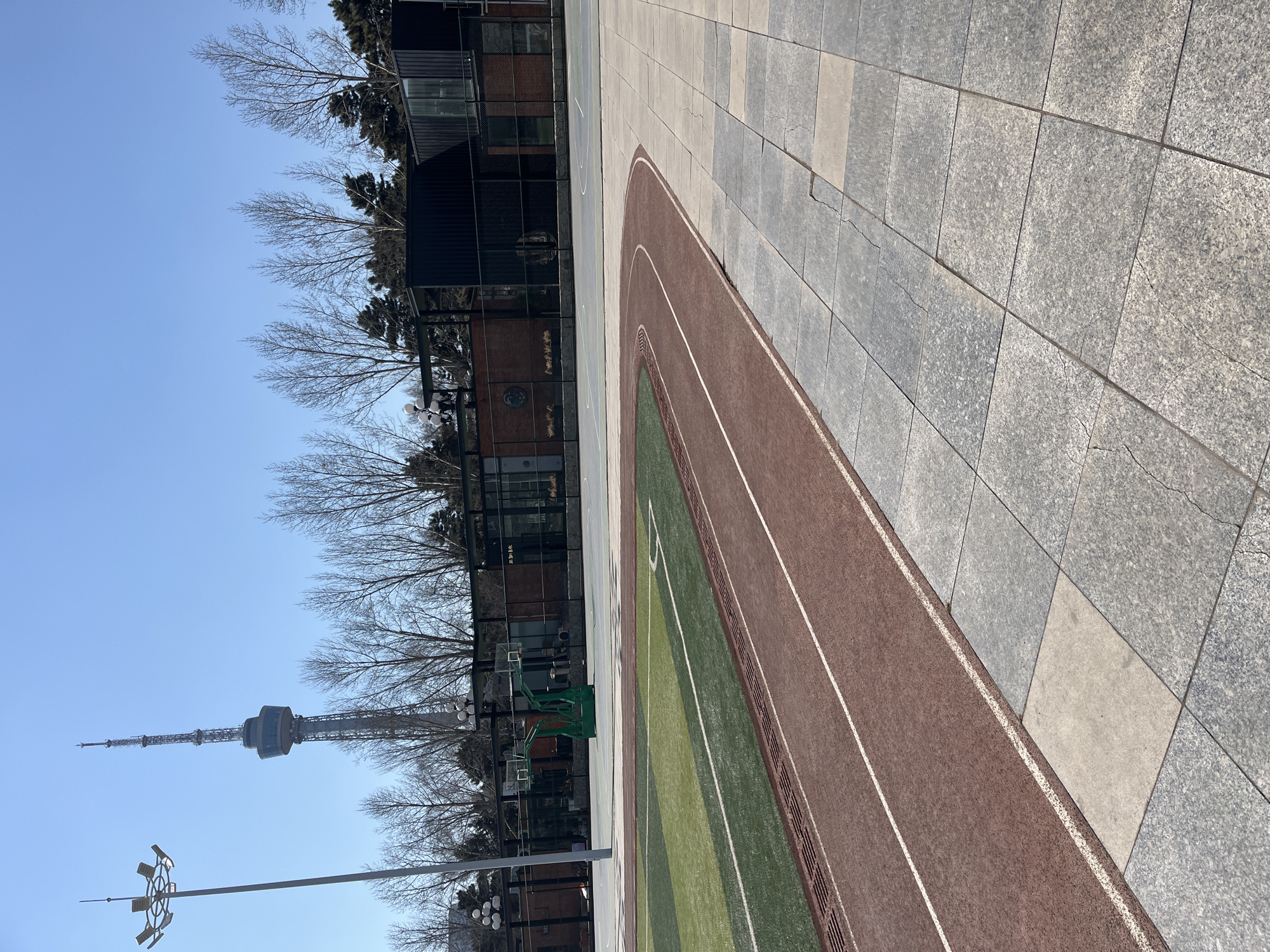 The image shows a track and field facility on a sunny day in Changchun City, China. A grey-tiled pedestrian area is adjacent to a red running track marked with white lines. A green sports field is within the track. A basketball court with green hoops is visible. A building with dark siding is situated next to the court. Bare trees stand between the court and the building, obscuring part of the view. In the background, a tall, slender television tower is visible against a clear blue sky. A light pole with multiple fixtures is also in view.