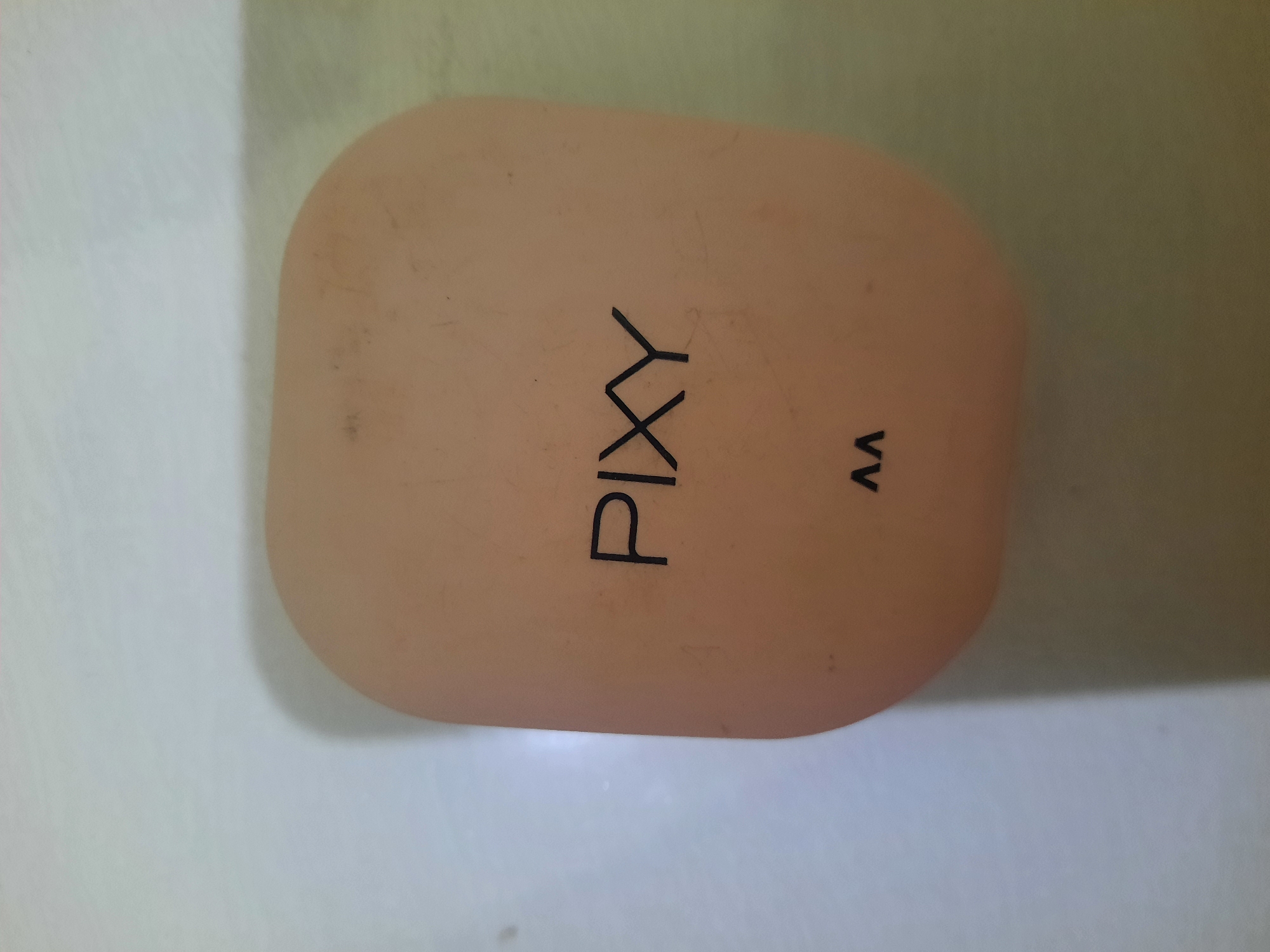 An overhead, close-up shot captures a light peach-colored, oval-shaped compact powder case from a high angle. The case has the brand name "PIXY" printed in black, sans-serif font across its surface, with a smaller "AA" logo below it. The surface of the case appears slightly worn with a few minor scuffs and dust marks. The background is a plain, light-colored surface, possibly a table or countertop, with soft shadows indicating overhead lighting. The lighting casts a subtle gradient across the background, with the right side appearing slightly darker and greener due to the reflected light from the object. The image composition focuses solely on the compact case, with no other objects or elements present in the frame. The location is not discernible from the image.