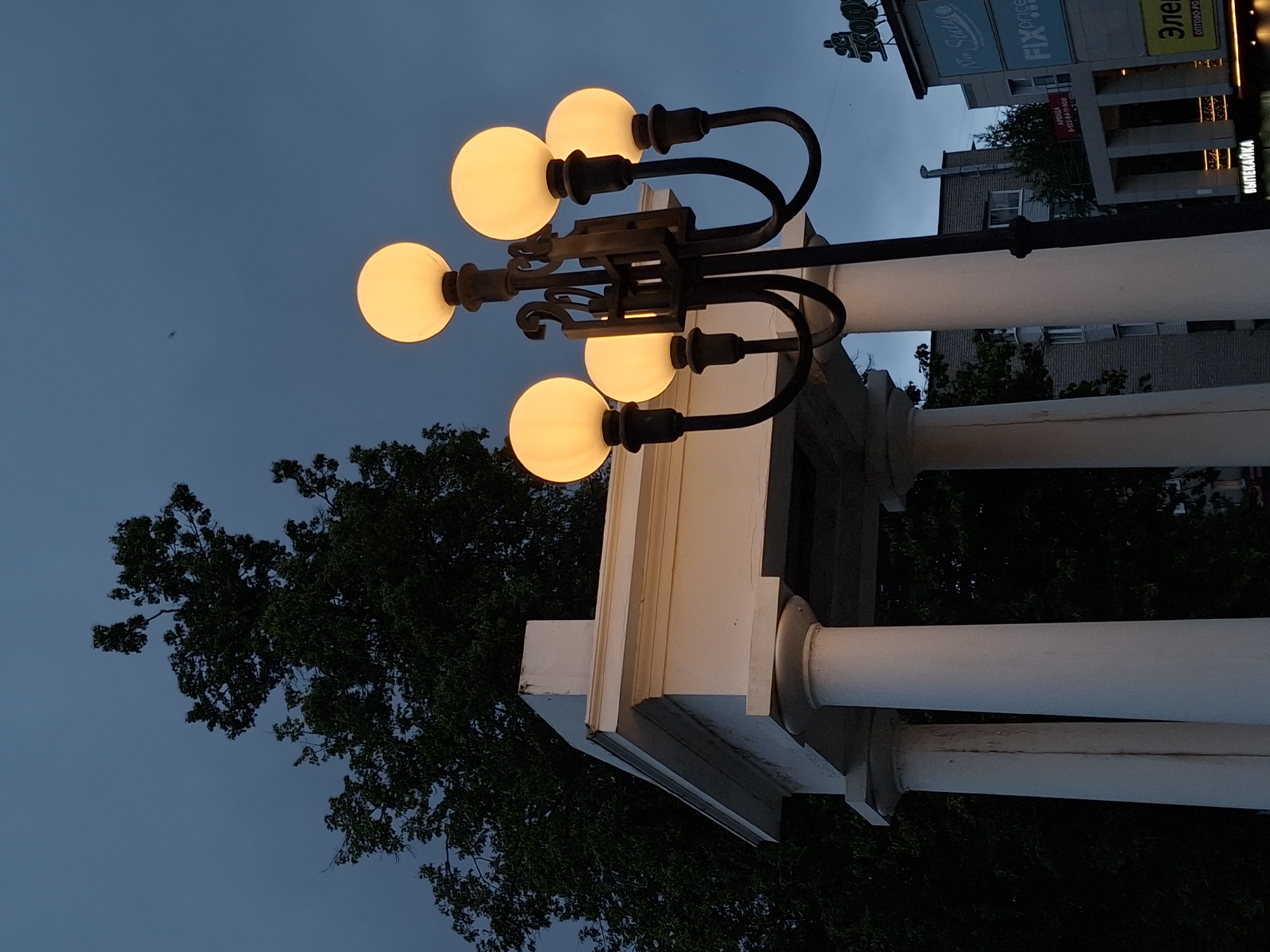 The image depicts an outdoor urban environment in Chaikovsky, Russia, at twilight or early evening, characterized by a dark blue sky. A prominent ornate lamppost, constructed of dark metal, stands on the right side of the frame, illuminating five spherical, amber-colored light fixtures. To the left and slightly behind the lamppost is a white classical-style structure, possibly a pavilion or portico, featuring at least two visible columns supporting an entablature. The leftmost column exhibits a distinct vertical crack. A large deciduous tree with dark foliage is silhouetted against the sky, positioned behind the white structure and extending into the upper left quadrant. A small, dark, indistinct object is faintly visible in the upper central sky area. In the background, several buildings are visible on the right. One building features a blue sign with "FIX price" and another partial sign reading "No Such". Below, a yellow and black sign displays "Элект" followed by "оптово-ро," and a smaller sign beneath it reads "ВЫПЕКАЙКА" (Bakery). An additional building exhibits a red "АРЕНДА" (FOR RENT) sign with a partial phone number. A partially obscured sign in the upper right background displays Cyrillic characters. No human figures are present.