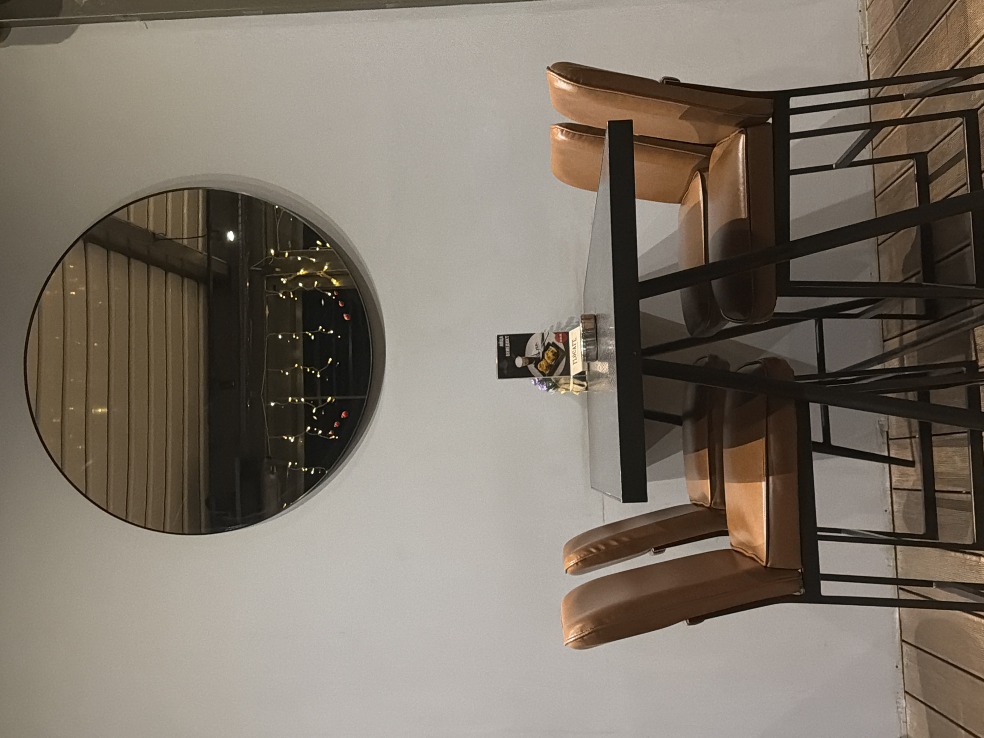 The image shows a brightly lit interior with a minimalist design. On the left, a circular mirror reflects wooden slats and indistinct decorative lighting. To the right is a table with a dark metal frame and what appears to be a glass top. Two chairs with brown leather seats are pulled up to the table. There is a promotional card on the table, and possibly a water bottle. No people are visible. There is no explicit information that places the location in Sofia, Bulgaria.