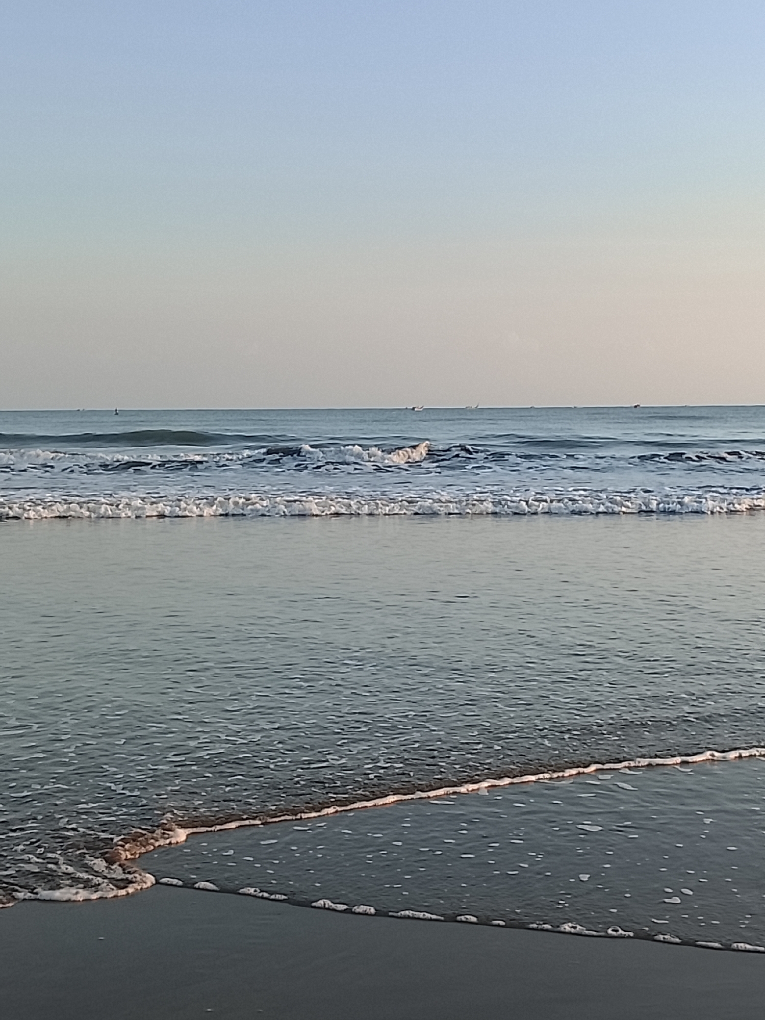 The image depicts a serene ocean scene at a beach in Reudeuep, Indonesia. The sky above is a soft gradient of pale blue, transitioning into a lighter hue near the horizon. The ocean itself is calm, with gentle waves rolling towards the shore. In the distance, several small boats are visible on the water. The foreground shows the wet sand of the beach, with small waves lapping at the edge and leaving a foamy trail. The overall impression is one of tranquility and natural beauty.