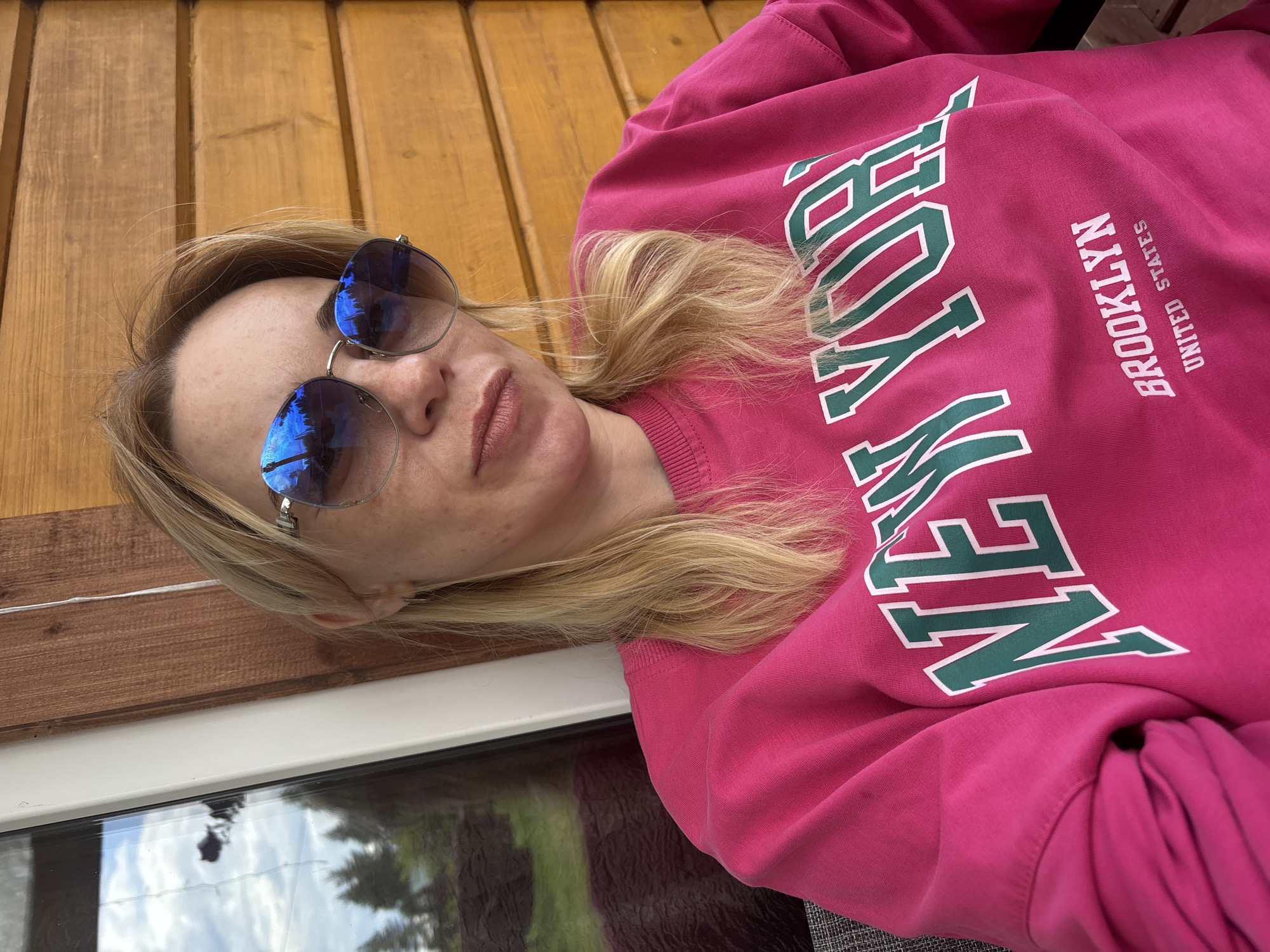 A woman with blonde hair and sunglasses is photographed outdoors, sitting in front of a wooden wall. The woman is wearing a bright pink sweatshirt with "NEW YORK" and "BROOKLYN UNITED STATES" printed on it in green and white letters. The background shows a window with a reflection of trees and sky. This image was taken in Shuiskaya Chupa, Russia.