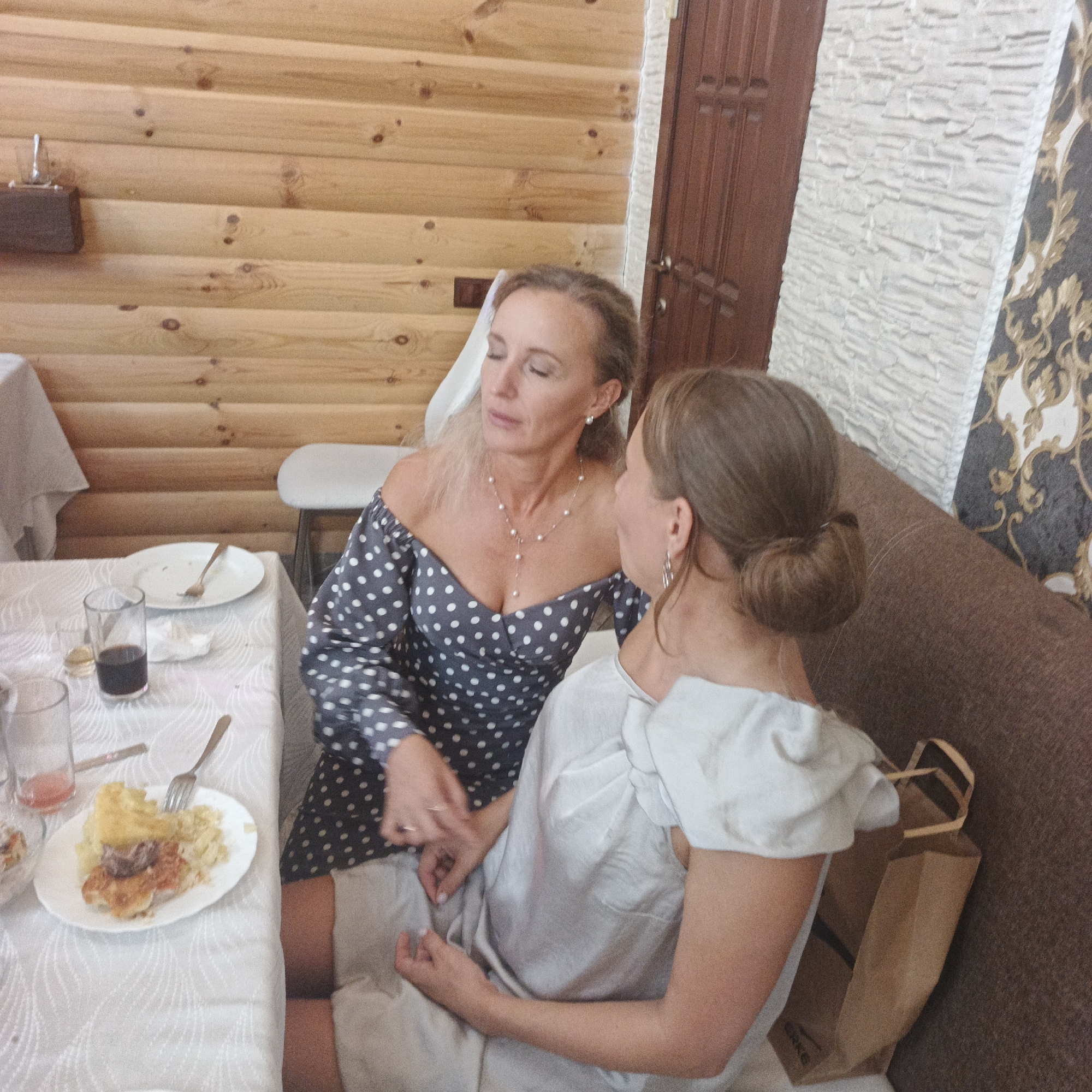 In the city of Chaikovsky, Russia, two women are seated at a table in what appears to be a rustic dining area. The room is decorated with a wooden plank wall. The woman on the left, wearing a dark blue polka dot dress with an off-the-shoulder neckline and a pearl necklace, is facing towards the right, with her eyes closed. The woman on the right, facing away from the camera and towards the left, wears a light-colored top. They appear to be in conversation or sharing a moment. On the table, partially visible, are two plates of food, cutlery, and glasses, suggesting a meal or a gathering. One plate contains what looks like mashed potatoes, meat, and some kind of pie or pastry. Another glass holds a dark liquid, possibly wine or juice. A brown paper bag is placed on the floor next to the woman on the right. The overall scene depicts a relaxed social setting.