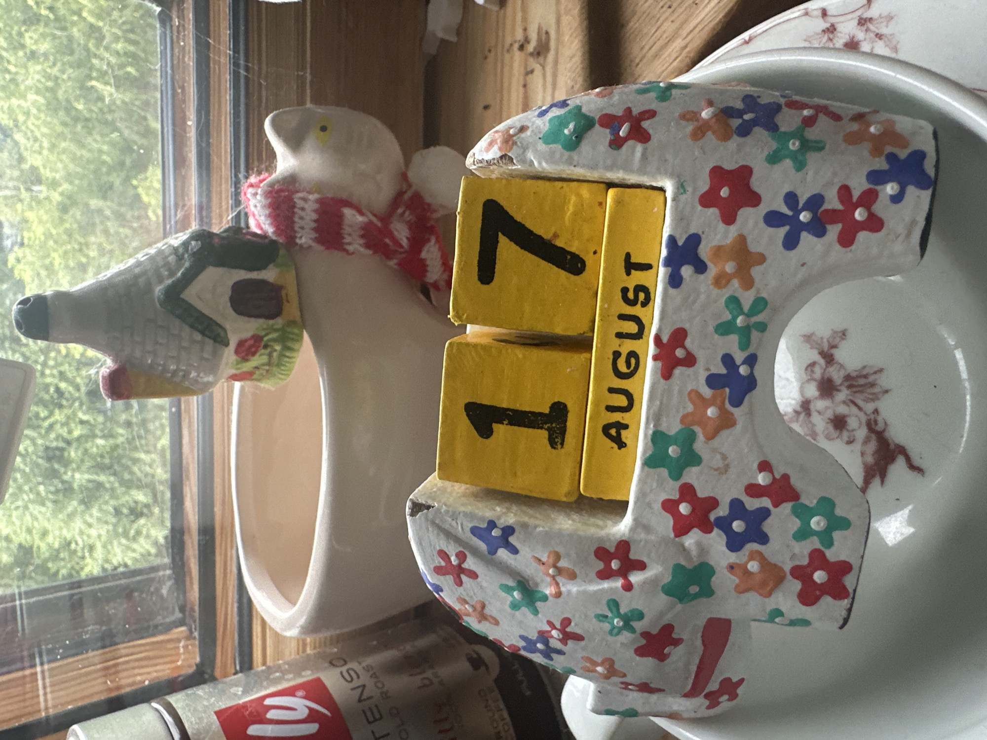 A still life photograph shows a decorative ceramic elephant displaying the date "17 AUGUST" on yellow blocks. The elephant is adorned with colorful floral patterns. Behind the elephant, a white ceramic pot and a miniature house-shaped object with a knitted scarf are visible. To the left, a metal tin with a red label, partially obscured, sits near a window. The background consists of a wooden surface and a view of green foliage through the window.