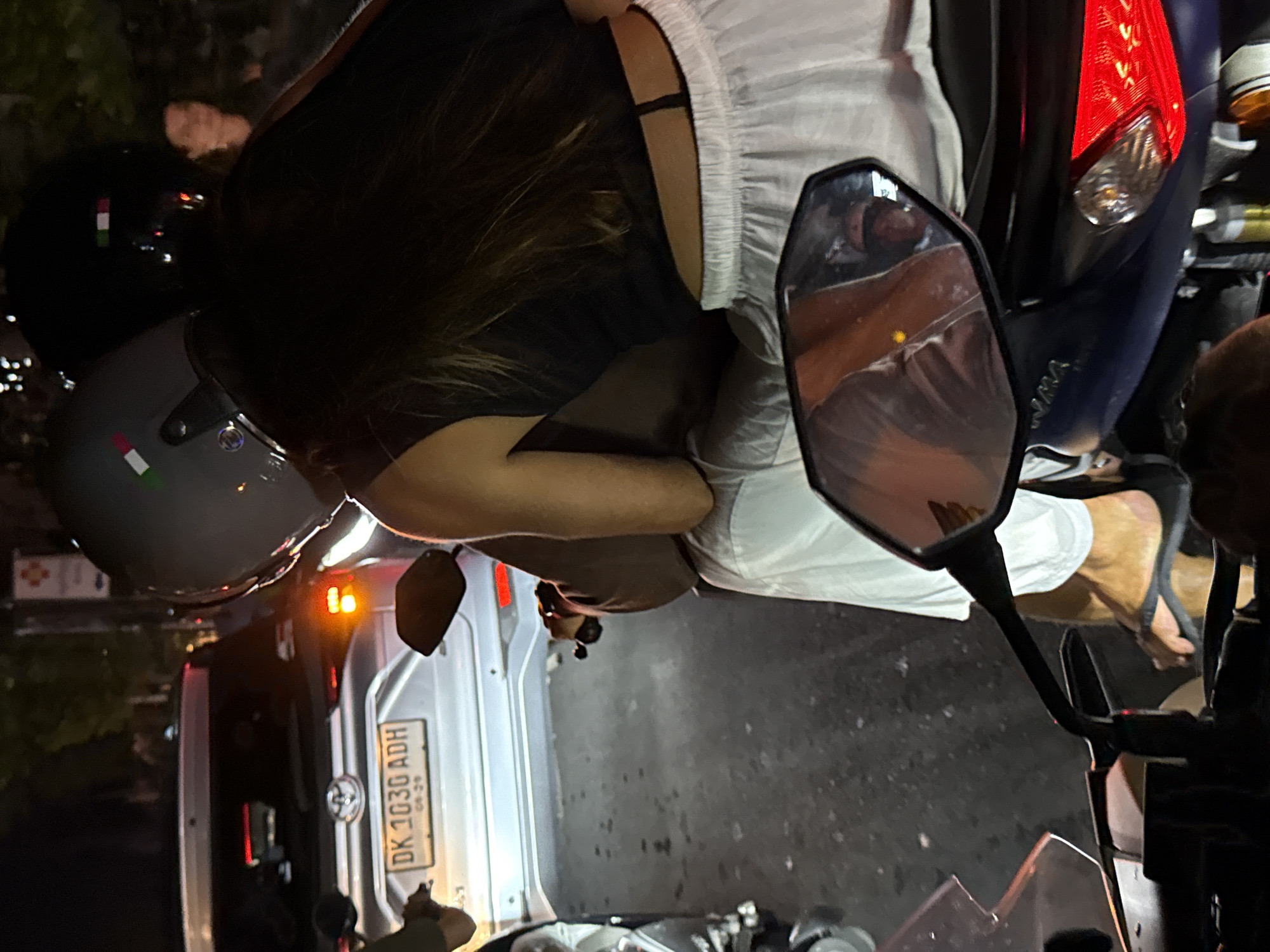 The image captures a nighttime street scene from the perspective of someone on a motorcycle or scooter, looking backward. In the immediate foreground, a portion of the motorcycle's handlebar and a rearview mirror are visible. The mirror reflects the upper body of a person, likely the rider, wearing a light-colored garment.
Seated on the motorcycle, with their back partially to the camera, is a person with dark hair, wearing a dark top and light-colored, possibly white, shorts or a skirt. Their right arm is visible, bent at the elbow. A gray helmet with a red, white, and green stripe (resembling the Italian flag) rests near them. Another black helmet is also visible to the left.
Behind the motorcycle, a light-colored car is stopped, possibly a Toyota, indicated by the emblem on its rear. The car's taillights are illuminated, and its right turn signal is blinking an amber light. A clear view of the car's license plate shows the text "DK 1030 ADH" and "09-29" below it, indicating a registration from Bali, Indonesia. The ground is dark asphalt, reflecting some of the vehicle lights. Distant, blurred lights suggest an urban or suburban setting. The scene depicts a moment of standstill, possibly in traffic or at an intersection, during the night in North Kuta, Indonesia.