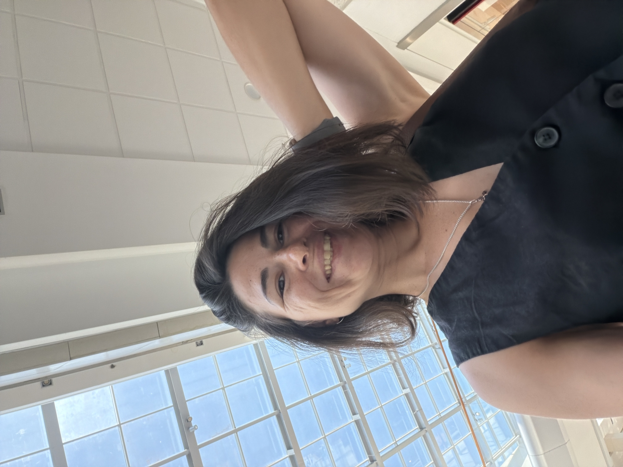 A woman with shoulder-length dark hair and a broad smile is looking up at the camera from a low angle. She is wearing a black sleeveless vest with two visible dark buttons on the front and a silver chain with a pendant. Her right arm is bent and raised, with her hand touching her head. The background features a modern ceiling with a glass-paneled skylight that allows natural light to illuminate the space, suggesting an indoor environment like a shopping mall or an atrium. The architectural style of the ceiling and the clean, bright lighting are indicative of a contemporary commercial or public building. The overall impression is a candid moment captured in a well-lit, open interior space. There are no identifiable landmarks that would suggest a specific location within Podgorica, Montenegro.