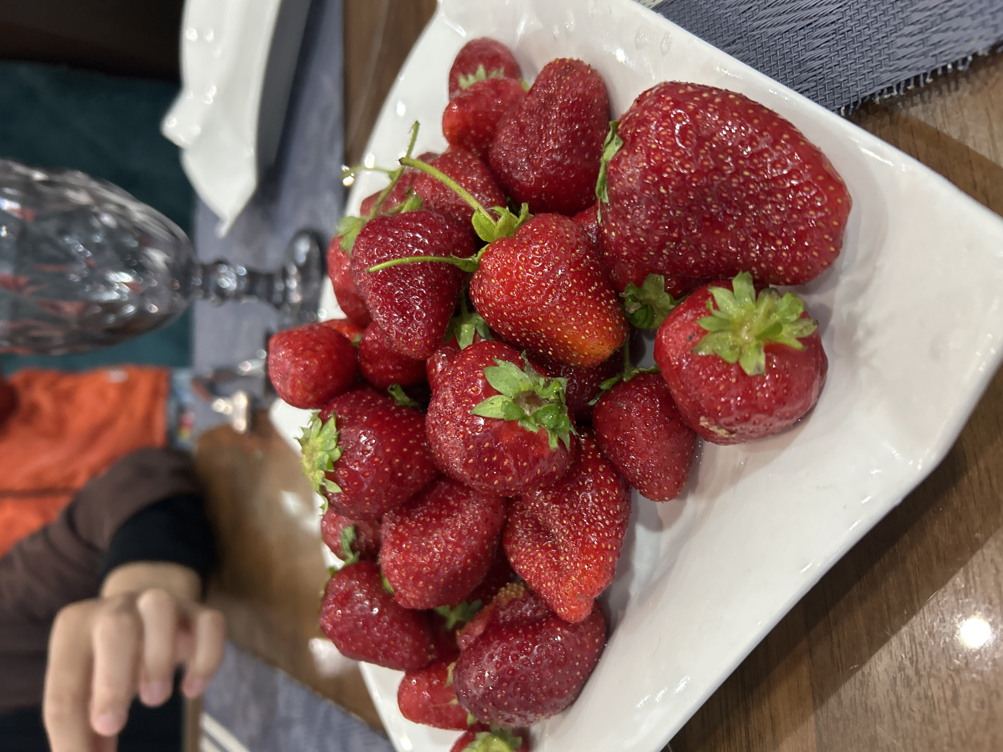 A close-up shot of a square white plate filled with fresh strawberries. The strawberries vary in size, with some having green stems attached. The plate is placed on a wooden surface with a blue and white patterned cloth visible. A hand is partly visible in the lower left corner, reaching towards the plate. A glass object, possibly a vase, is blurred in the background on the left side of the image. The photo does not provide information about the city of Ақсу, Kazakhstan.