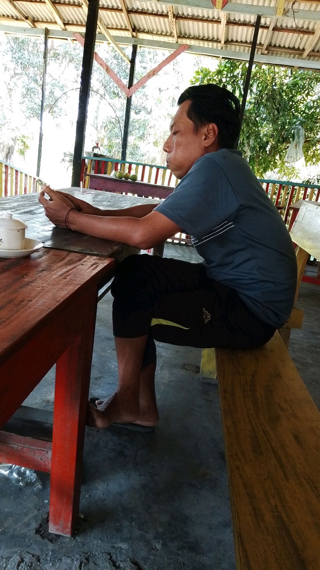A man is sitting on a wooden bench on the left side of the image, facing away from the viewer and looking to the left. He is wearing a dark blue t-shirt and black pants with a yellow stripe. His arms are resting on a wooden table. He is holding a smartphone in his left hand. On the table, there is a white cup and saucer. The man is in a covered outdoor seating area with a corrugated metal roof. The area appears to be a cafe or restaurant in Mojopetung, Indonesia, given the style of the seating and the surrounding tropical vegetation.