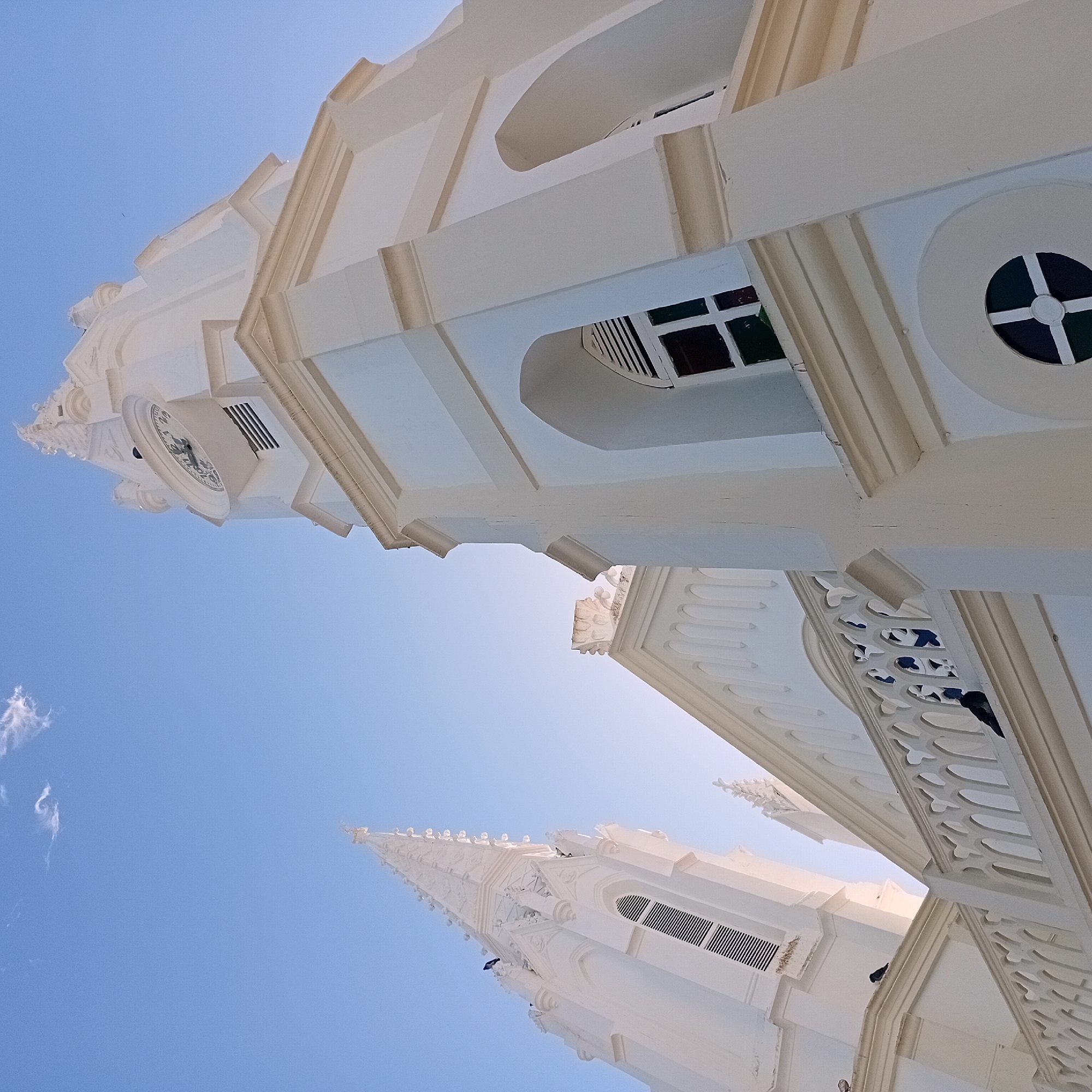 The image shows the exterior of a white church. The architecture includes towers, a clock, and arched windows with colored glass. The sky is blue and there are a few wisps of white clouds visible. The image is taken from a low angle, looking upwards towards the church spires. The exact location is unknown.
