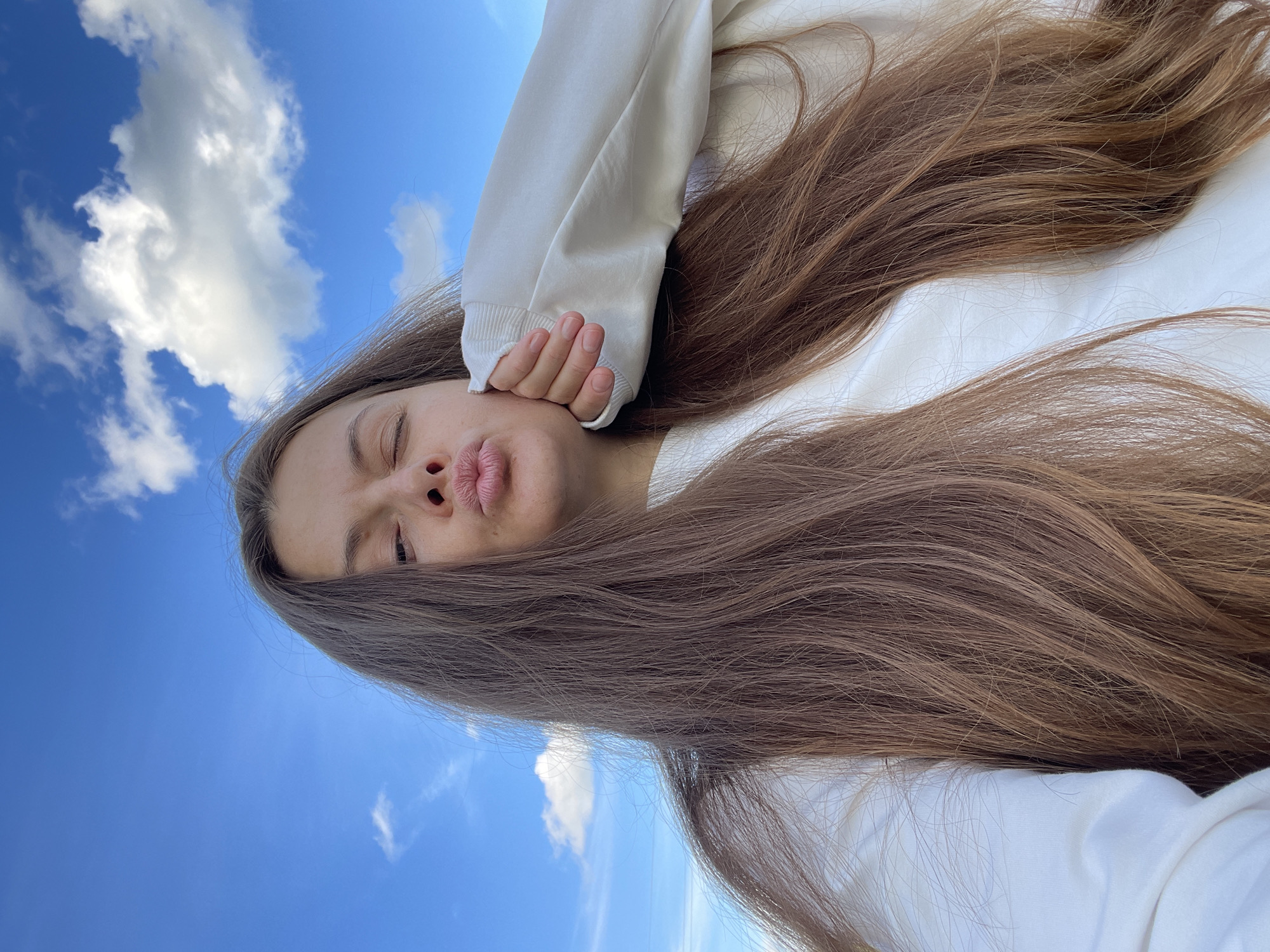 A young woman with long, brown hair is captured in a selfie with a bright blue sky and scattered white clouds as the backdrop. She is wearing a white sweatshirt and is puckering her lips while resting her chin on her hand. The image appears to have been taken outdoors.