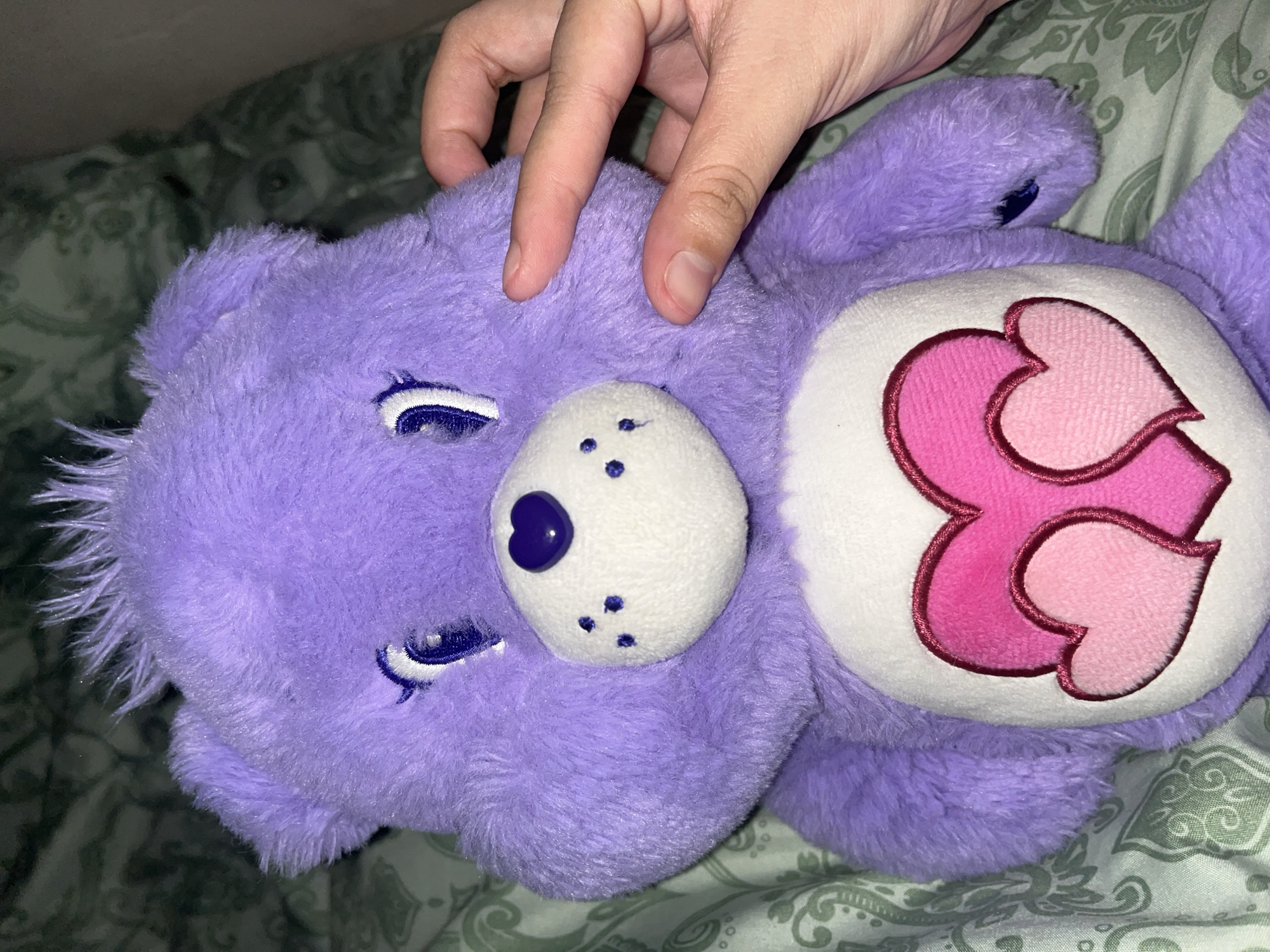 The image displays a purple plush stuffed bear, identified as a Care Bear, with fuzzy texture and spiky fur on its head. It has embroidered white eyes with blue pupils and white highlights. Its white snout features a prominent purple heart-shaped nose and three small blue embroidered dots on each side. On its chest is a white circular patch with an embroidered design of two overlapping pink hearts, one lighter and one darker. A human hand, with light skin tone and short, clean fingernails, is positioned on the bear's proper right side, gently pressing against its face and fur. The background consists of a green fabric surface adorned with a repeating pattern of light green, intricate, swirling designs, suggestive of bedding. The scene is set in an indoor environment. No discernable urban or exterior geographical features are present in the image to identify a city or specific outdoor location.