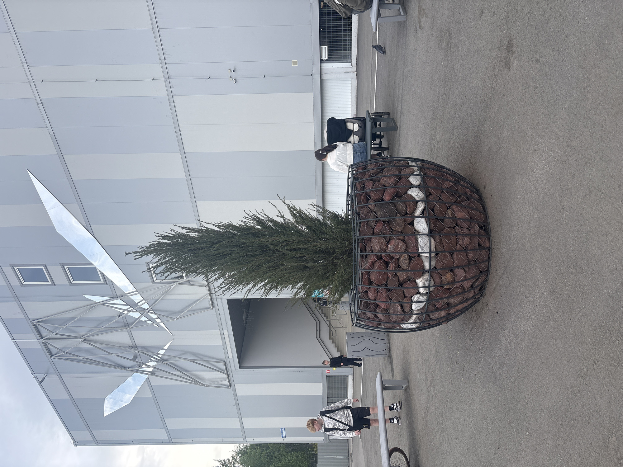The image shows an outdoor scene in Perm, Russia. A large, spherical wire basket filled with red and white rocks contains a coniferous tree. This arrangement sits on a gray concrete surface. Behind the tree is a modern building with a gray and white striped facade and an architectural feature resembling reflective wings above the entrance.
Several people are visible. One person is seated on a bench near the tree. Another person is walking towards the building entrance, while a third person, possibly a child, stands near a bicycle at the bottom of the frame. The environment appears to be a plaza or pedestrian area outside the building. The sky is overcast.