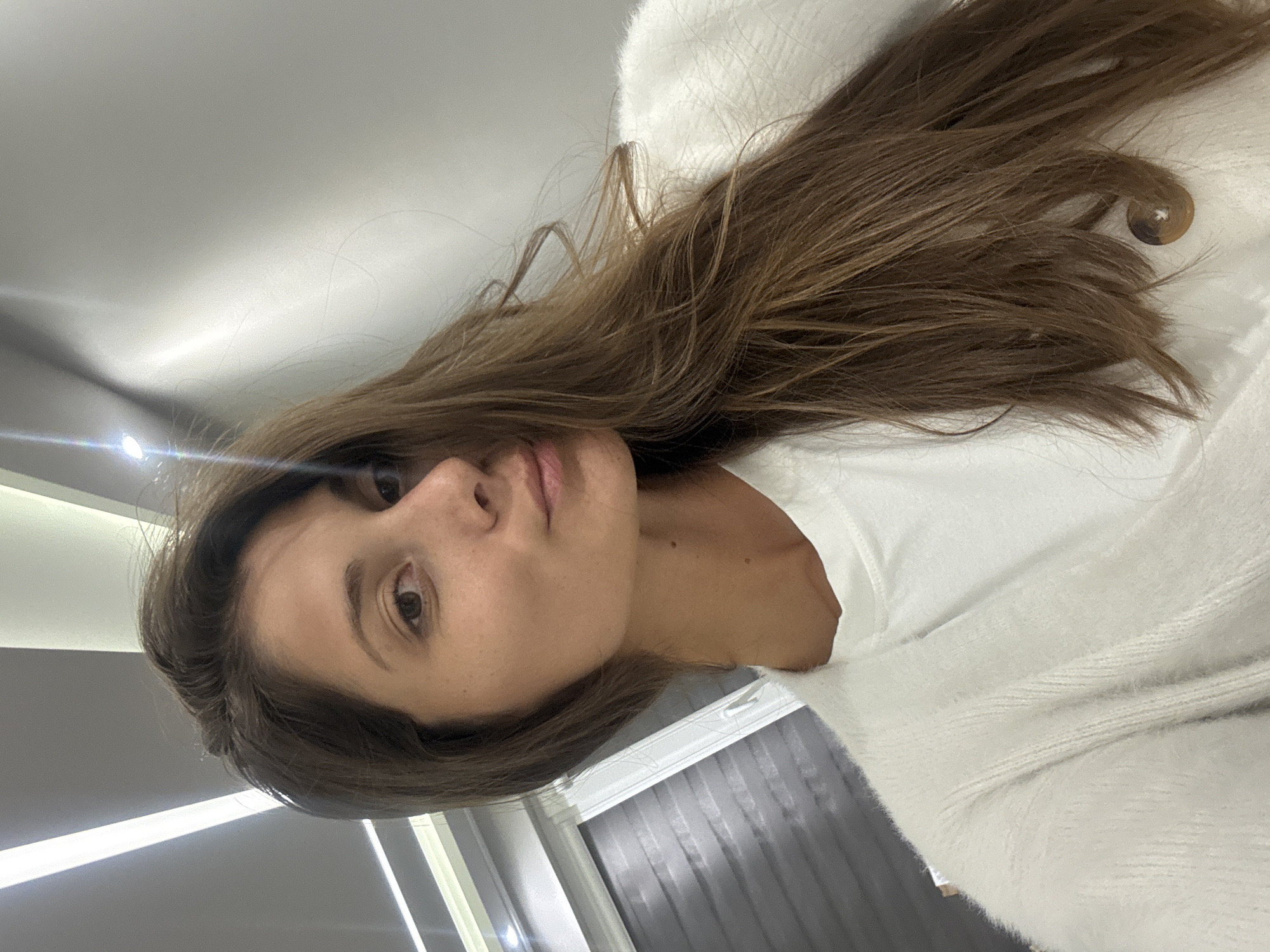 A woman with long brown hair is indoors, wearing a white top and a fuzzy white sweater. She is looking at the camera. She is inside a room with light-colored walls, a window with grey blinds, and a light fixture on the ceiling. There is no information available about the location being in Burgas, Bulgaria.