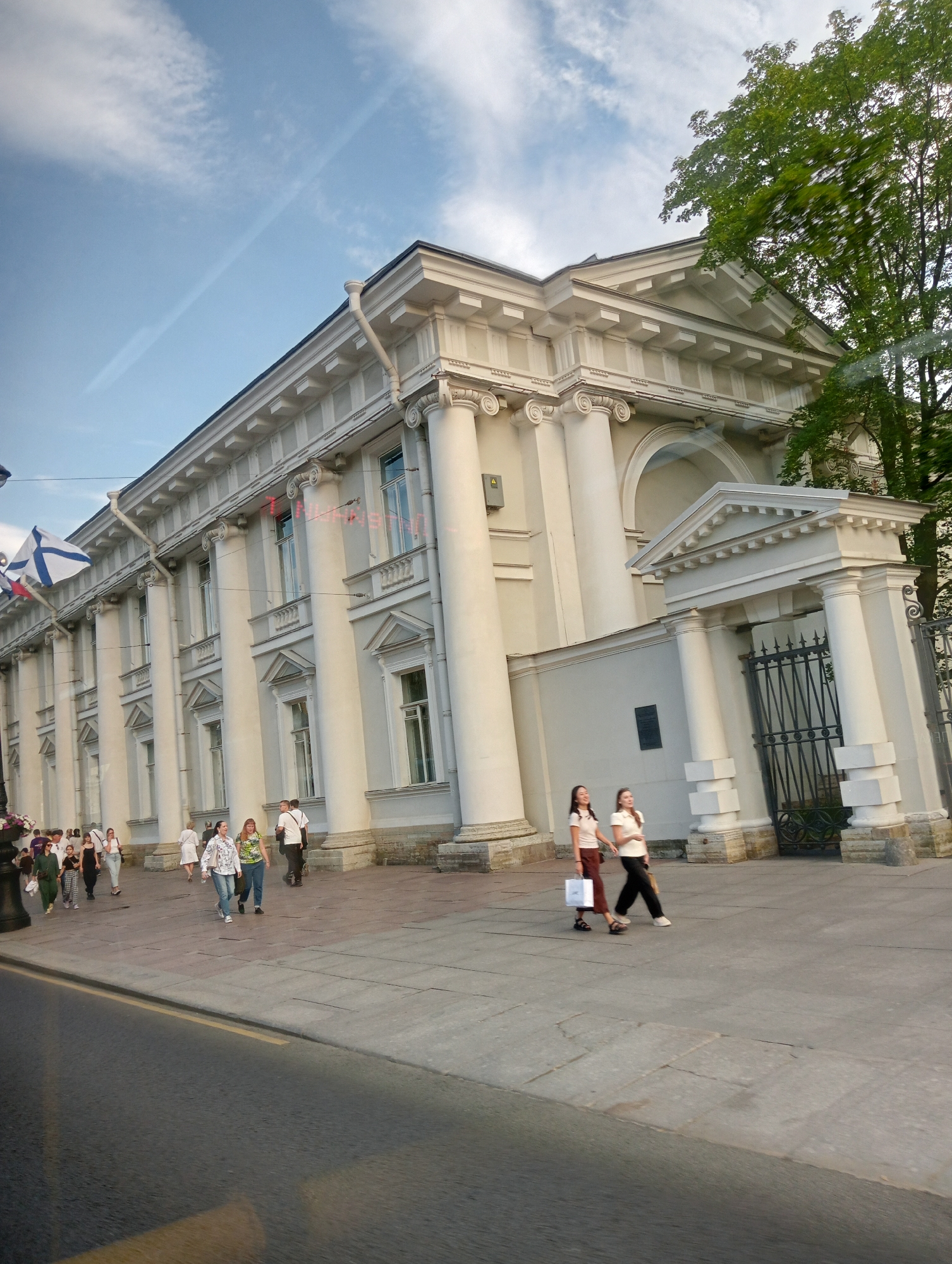 The image depicts the facade of a classical building in Saint Petersburg, Russia, likely an administrative or cultural institution given its grand architecture. The building is characterized by a series of large Corinthian columns supporting a prominent entablature and pediment. Several arched windows are visible along the facade, some featuring decorative elements. To the left, a flag with a blue cross on a white background is flying.
In the foreground, on the paved walkway in front of the building, several individuals are walking. On the right side, two young women are walking towards the right. The woman on the left wears a light-colored shirt and a brown skirt, carrying a white shopping bag. The woman on the right wears a white polo shirt and dark pants. Further to the left, a group of people are seen walking, including a man in a white shirt and dark pants, and several women in casual attire. Some appear to be tourists.
The environment suggests a daytime scene with a partly cloudy sky. A large green tree with dense foliage is visible on the far right, partially obscuring the building's upper section. A black wrought-iron gate is present to the right of the building's entrance, leading into an area behind a wall. The street in the lower part of the image appears to be asphalt, with a yellow line marking its edge. The overall impression is one of a public space with a mix of architecture and pedestrian activity.