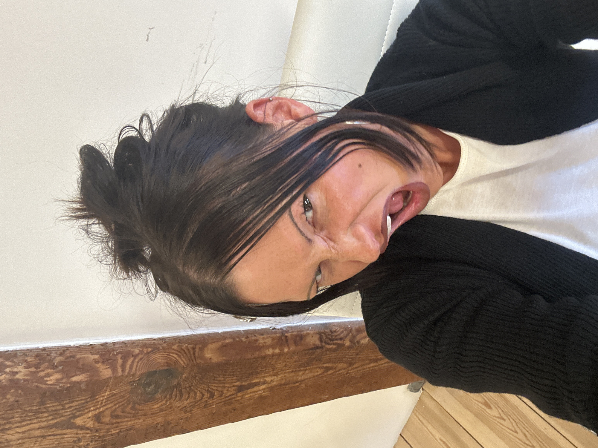 A sideways portrait captures an individual with dark hair and an exaggerated facial expression, taken indoors. The person is seen from the chest up, wearing a white t-shirt beneath a black ribbed cardigan. Their head is tilted sharply, creating a distorted appearance, with the left side of the face prominent. Dark hair is tied up, but several strands fall across the forehead and cheek. A silver stud piercing is visible in the upper cartilage of the left ear. One eye is wide open, featuring what appears to be dark eyeliner, and the mouth is open in a broad, expressive manner, revealing the tongue. The expression suggests a playful or theatrical pose.
The background features a plain white wall above a prominent, horizontal wooden beam or railing. This wooden element displays a distinct grain, knots, and signs of wear, indicating a rustic or natural aesthetic. A light-colored wooden floor is partially visible in the lower portion of the scene. The lighting appears even and diffused, characteristic of an indoor setting, likely in London, United Kingdom. No text is visible in the image.