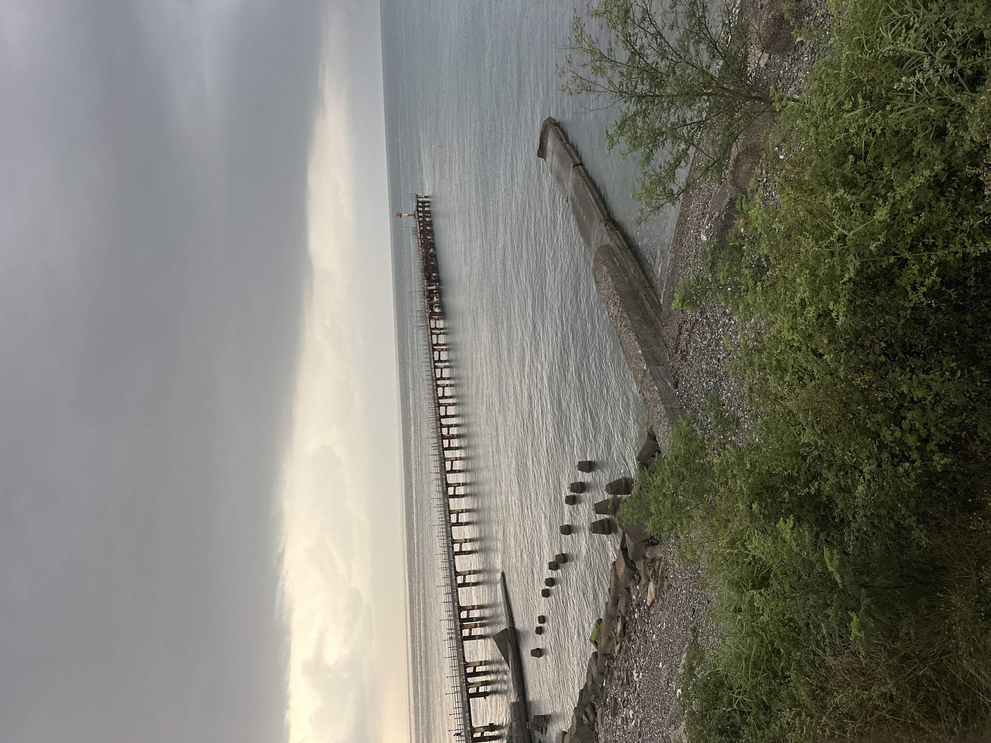 An overcast day at an unknown location by the sea. A long pier extends out into the water, supported by numerous pilings. A small lighthouse with red and white markings stands at the end of the pier. The water is calm, with a slight ripple. Along the shoreline, there are remnants of concrete structures and a rocky beach. Green bushes and trees are visible in the foreground.
