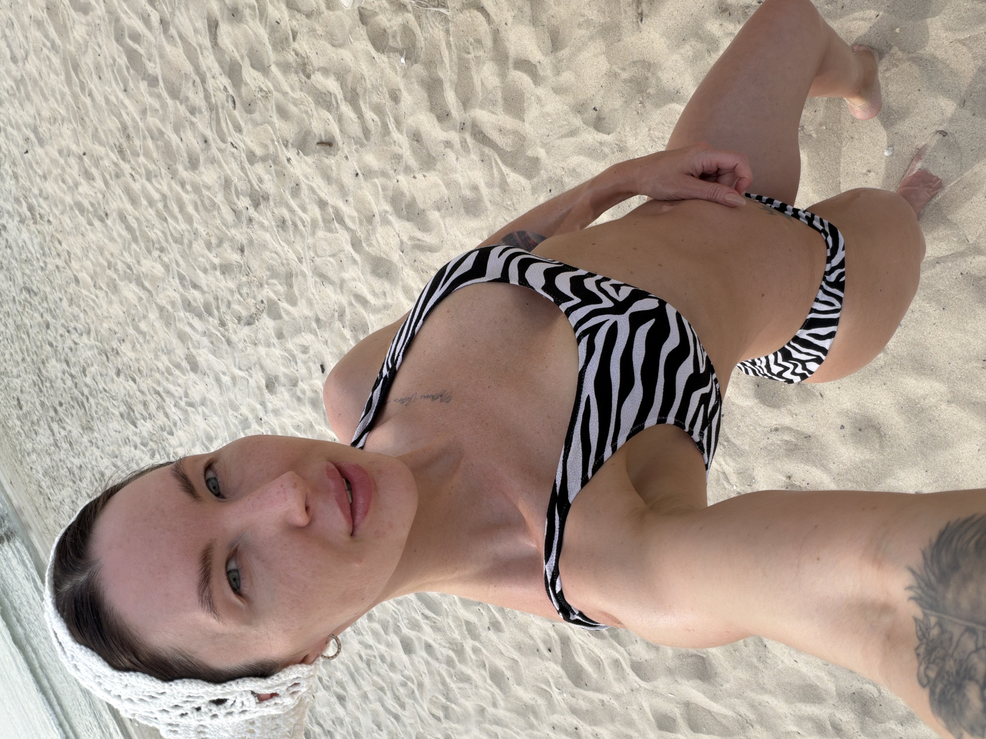 A woman wearing a white crocheted headscarf and a black and white zebra-print bikini poses for a selfie on a sandy beach. The woman has light skin, light eyes, and light brown hair. She is looking at the camera with a slight smile. The background is a sandy beach with gentle waves in the distance. The location is a beach in Dong Ninh Hoa Ward, Vietnam.