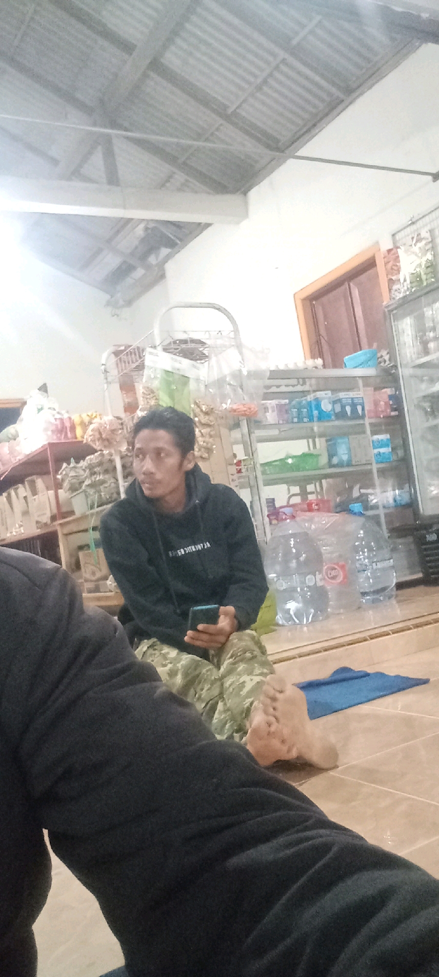 The image depicts a young man sitting on the floor in what appears to be a small shop or storage area. He is wearing a black hooded sweatshirt and camouflage pants. He is holding a smartphone in his right hand and looking towards it. In the background, there are shelves stocked with various goods, including bottled water and packaged items. The ceiling is corrugated metal, supported by wooden beams. The floor is tiled. The overall setting suggests a humble commercial space, possibly in Kras, Indonesia, given the user's prompt. The lighting is dim, indicating it might be evening or an indoor space with limited light sources.