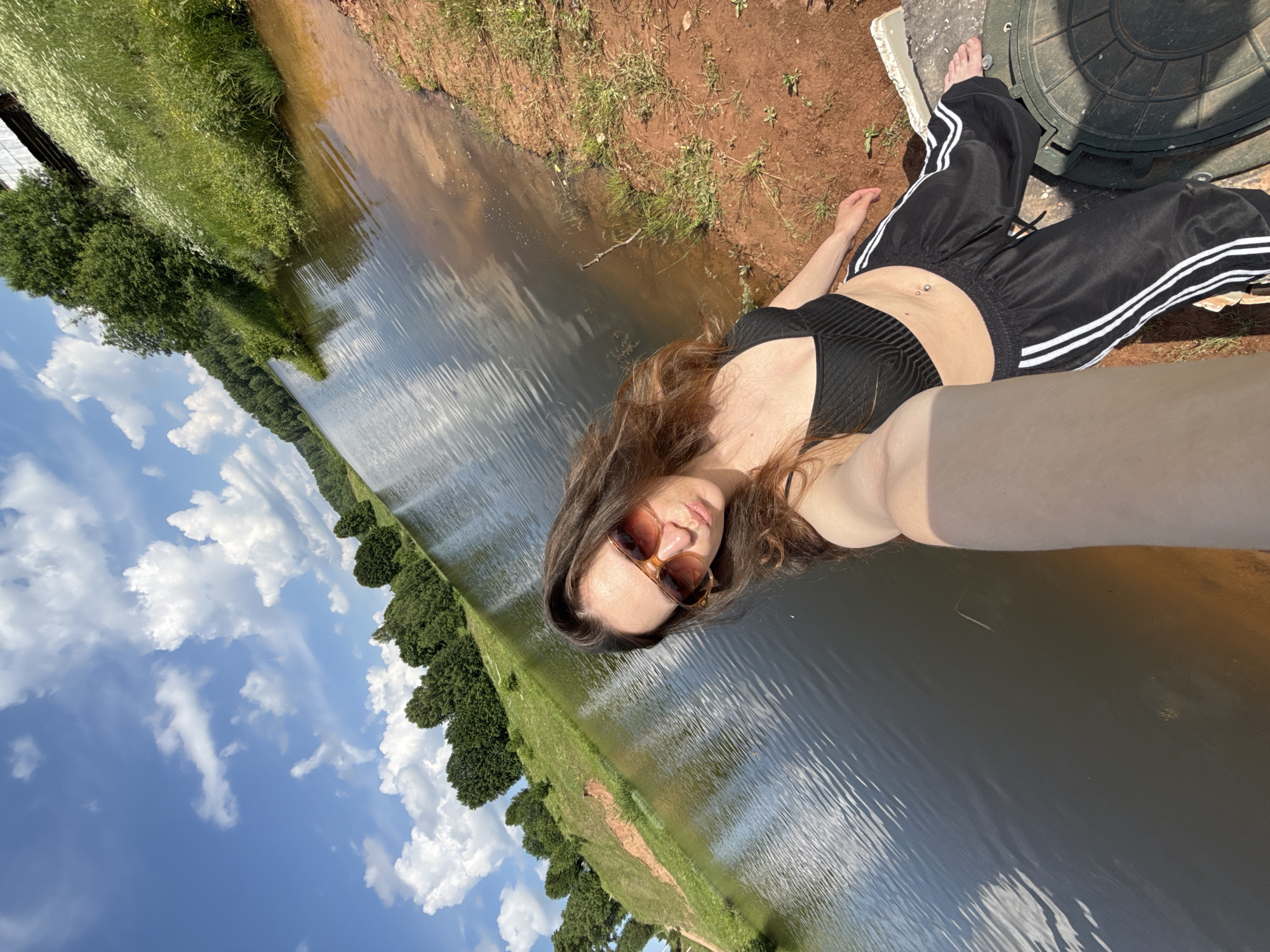 A young woman with long, dark, wavy hair and sunglasses is taking a selfie on a sunny day by a body of water. She is wearing a black sports bra and black pants with white stripes. The background shows a lake, a grassy embankment with trees, and a blue sky with white clouds. The ground she is sitting on is reddish-brown soil. The image is taken from a high angle, looking down at the woman and the scenery.