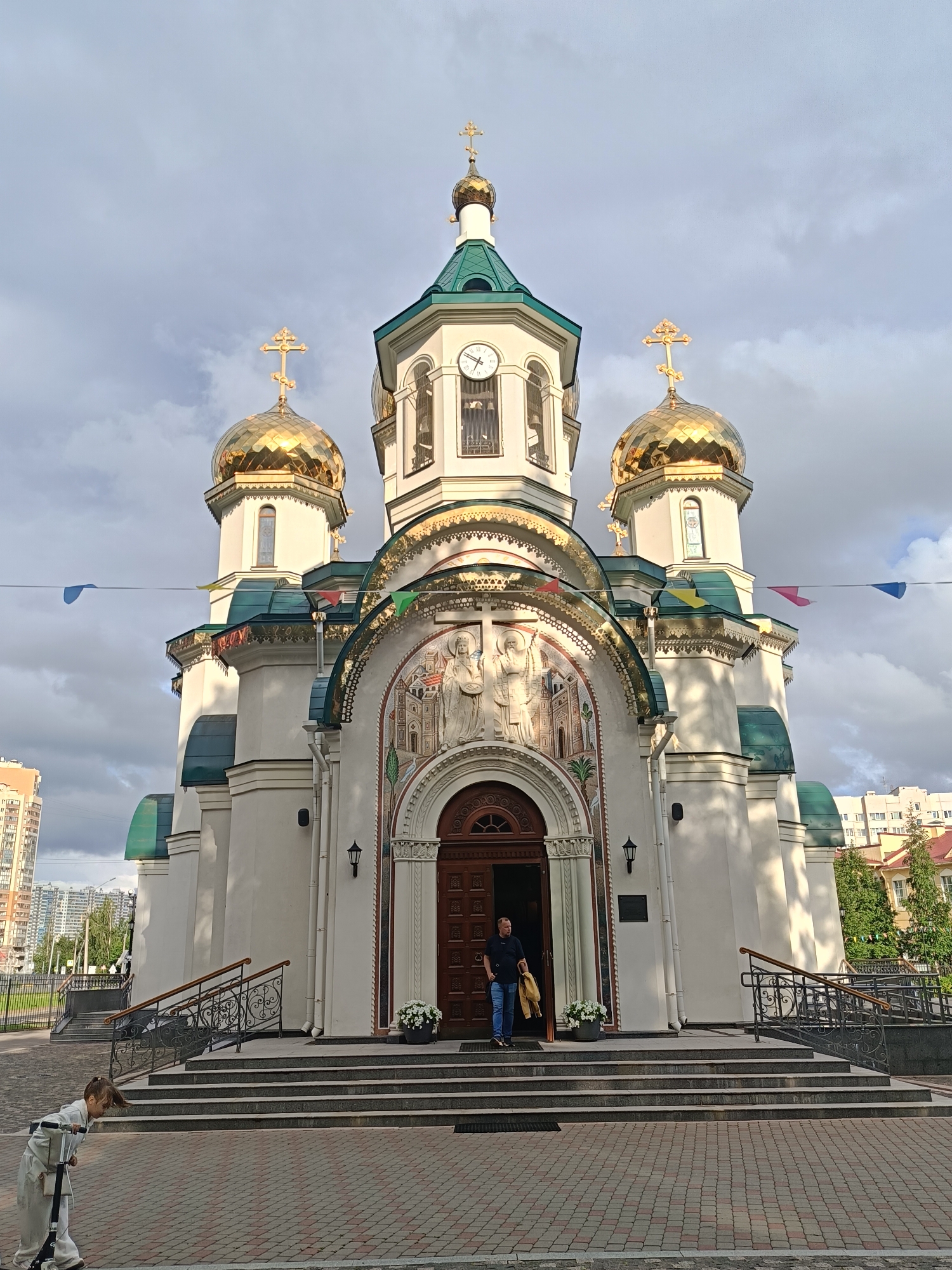 The image shows the Church of the Icon of the Mother of God "Joy of All Who Sorrow" in Saint Petersburg, Russia. The church features white walls, a green roof, and golden domes topped with crosses. A man stands in the open doorway, holding a yellow jacket. A young girl with a scooter is visible in the lower left corner. The church is located in a modern urban environment, with apartment buildings visible in the background. The sky is cloudy.