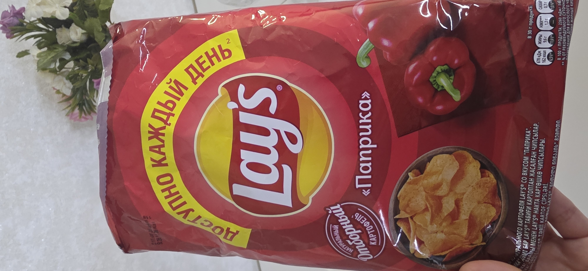 The image shows a close-up of a bag of Lay's paprika-flavored potato chips. The bag is primarily red with a yellow curved band containing the text "ДОСТУПНО КАЖДЫЙ ДЕНЬ" in yellow Cyrillic letters. The Lay's logo is prominently displayed in the center. To the left of the logo, there is a circular emblem with text that appears to be in Russian, indicating "НАТУРАЛЬНЫЙ ОТБОРНЫЙ КАРТОФЕЛЬ" (Natural Selected Potato). Below this, a bowl of potato chips is depicted. To the right of the logo, the word "Паприка" (Paprika) is written in quotation marks. Two red bell peppers are also visible on the right side of the bag. Nutritional information is displayed in the lower right corner. The overall presentation is bright and vibrant, typical of food packaging. There are no visible landmarks or indications of the city of Sumgayit, Azerbaijan in the image.