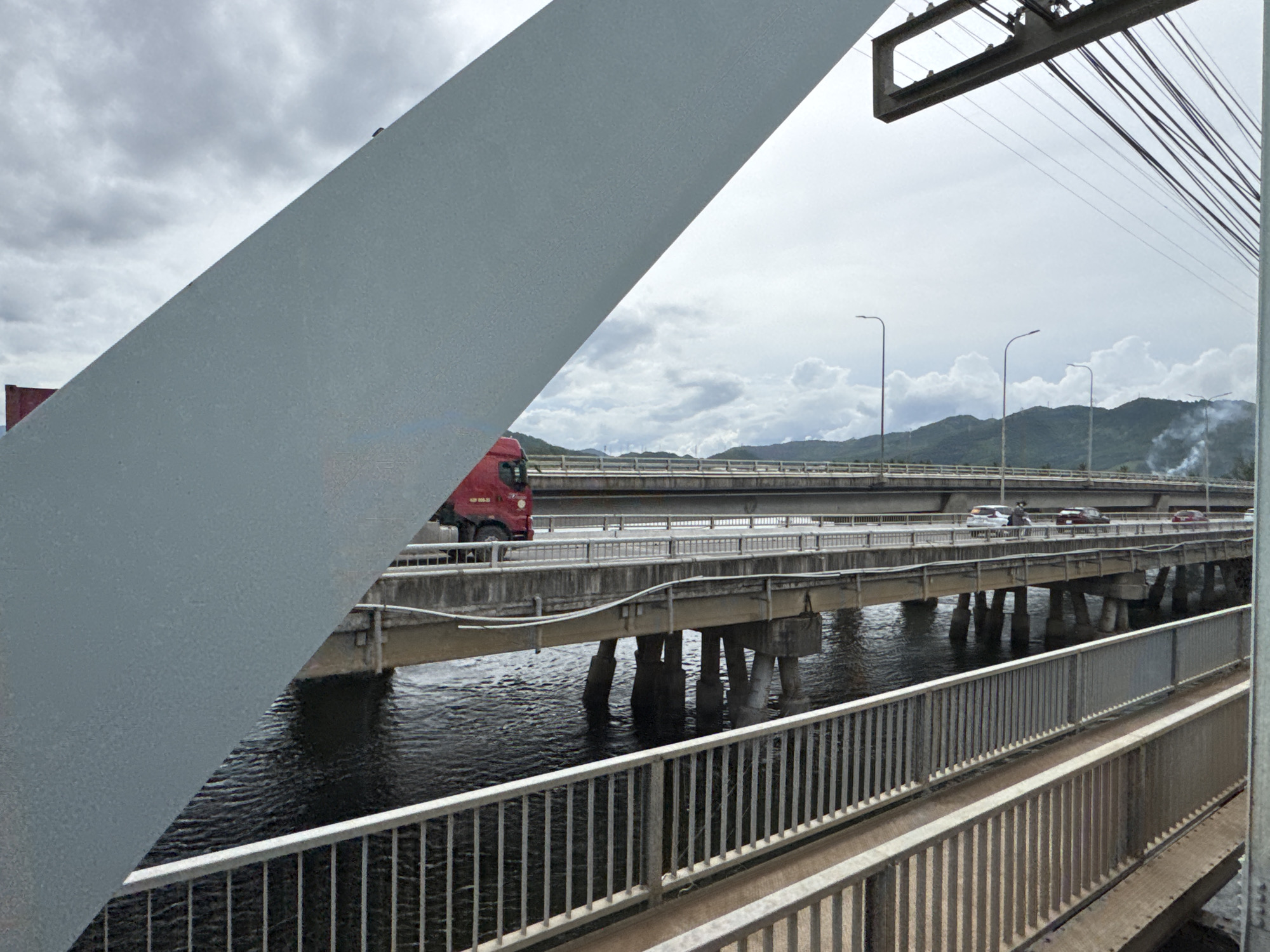 This image depicts a view of the bridges and traffic in the city of Đà Nẵng, Vietnam. In the foreground, a large, gray, diagonal structural element of a bridge dominates the left side of the frame, obscuring a portion of the scene. To the right, a red truck is visible on a raised highway, moving from left to right. Beyond this, multiple parallel elevated roadways span a body of water. Several cars are seen driving on these bridges. The background features a hazy sky with scattered clouds, and in the distance, verdant, hilly terrain with plumes of smoke suggests industrial activity or natural phenomena. Streetlights and overhead electrical wires are also present, indicating an urban or semi-urban environment. The overall atmosphere is overcast.