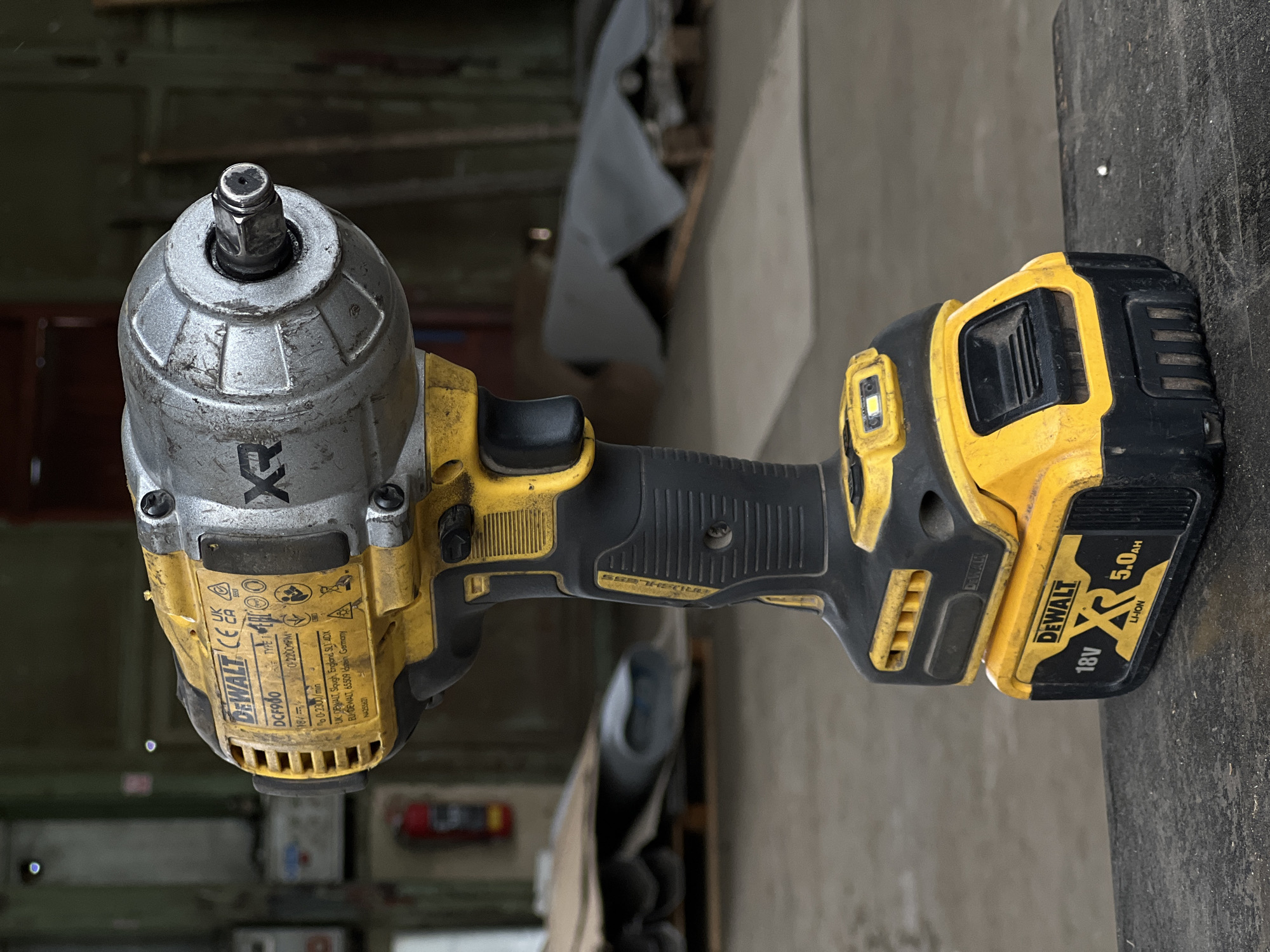 This is a close-up, rotated photograph of a DeWalt DCF980 cordless impact wrench, likely taken in a workshop or garage setting. The tool is primarily yellow and black, with a silver metal housing around the anvil. Visible text on the tool includes branding like "DEWALT", "XR", and "BRUSHLESS," along with model and technical specifications such as "DCF980," "18V," and "5.0Ah." There are also certification marks like "CE" and "UKCA." The impact wrench is not actively in use, but its presence suggests a context of construction, repair, or automotive work. The background is out of focus, showing hints of shelving, materials, and a concrete floor, reinforcing the workshop environment. The lighting appears to be artificial or overcast, with no strong shadows suggesting a specific time of day. The overall impression is that of a tool ready for use in a utilitarian setting.