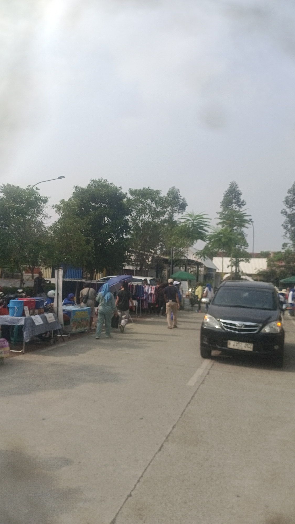 A street market scene is captured on a cloudy day in Tangerang, Indonesia. Several stalls are set up along the roadside, displaying various goods, including clothing. People are seen browsing the stalls and walking along the street. A black car is parked on the right side of the image, and a motorbike is partially visible on the left. The environment appears to be an outdoor public space with trees lining the background.