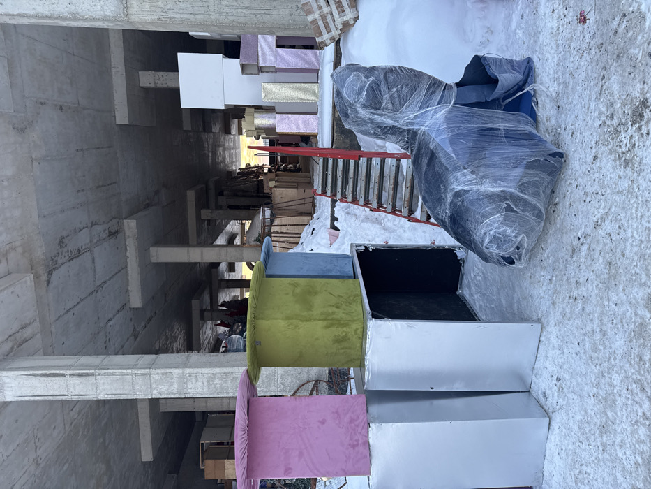 The image displays a winter scene in L'vovskiy, Russia, situated on snow-covered ground adjacent to or beneath an unfinished concrete structure. The structure features exposed concrete pillars and a flat, unpainted concrete ceiling.
In the foreground, from left to right, three square-topped pedestals or tables, upholstered in velvet-like fabric, are positioned on top of silver-colored, rectangular box-like units. The pedestals are pink, lime green, and light blue. One silver box beneath the blue pedestal is open, revealing a dark interior. To the right, a dark blue armchair, completely encased in transparent plastic shrink wrap, rests directly on the snow.
In the mid-ground, a temporary staircase with red metal frames and wooden steps descends into the concrete structure; snow is visible on and around the steps.
The background, within the confines of the concrete structure, contains various materials. To the left, wooden pallets and other construction supplies are stacked. To the right, a stack of rectangular boxes, some white, others covered in pink or gold glitter, are arranged against a concrete pillar. Distant, indistinct shapes or figures are visible deeper within the structure, illuminated by ambient light from an unseen opening. No immediate human activity is observed in the primary view.