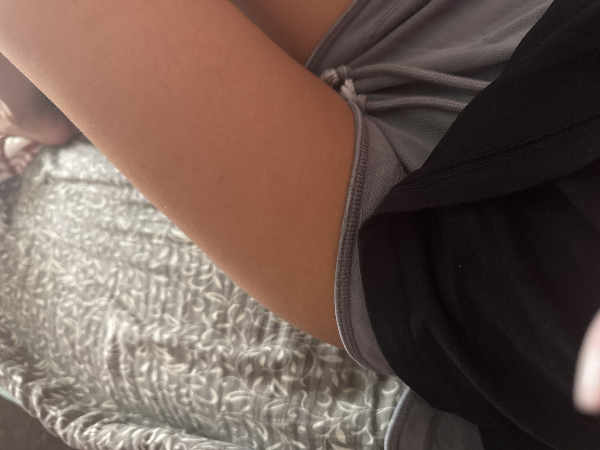 The image shows a close-up view of a person's legs and lap, lying on a bed with a grey patterned duvet. The person is wearing black shorts and grey athletic bottoms. The lighting suggests an indoor setting, likely a bedroom. There is no discernible indication of the city of Basarabeasca, Moldova, or any other specific location. The focus is on the clothing and skin, with the background out of focus.