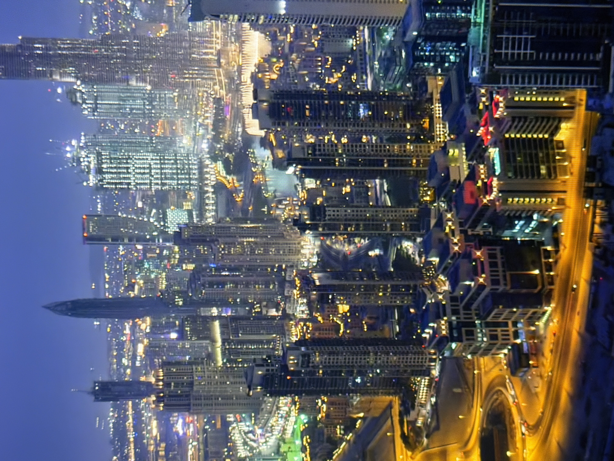 This is an aerial, night-time photograph of a densely packed urban skyline with numerous illuminated skyscrapers. The buildings display a variety of architectural styles and heights, with many featuring intricate lighting patterns. Several of the skyscrapers are under construction, indicated by visible scaffolding and cranes. The city is situated near a body of water, possibly a river or a bay, which reflects the city lights. The overall impression is that of a modern, developed metropolitan area. The scene is devoid of any identifiable individuals or specific landmarks that would pinpoint its location to Kyiv, Ukraine.