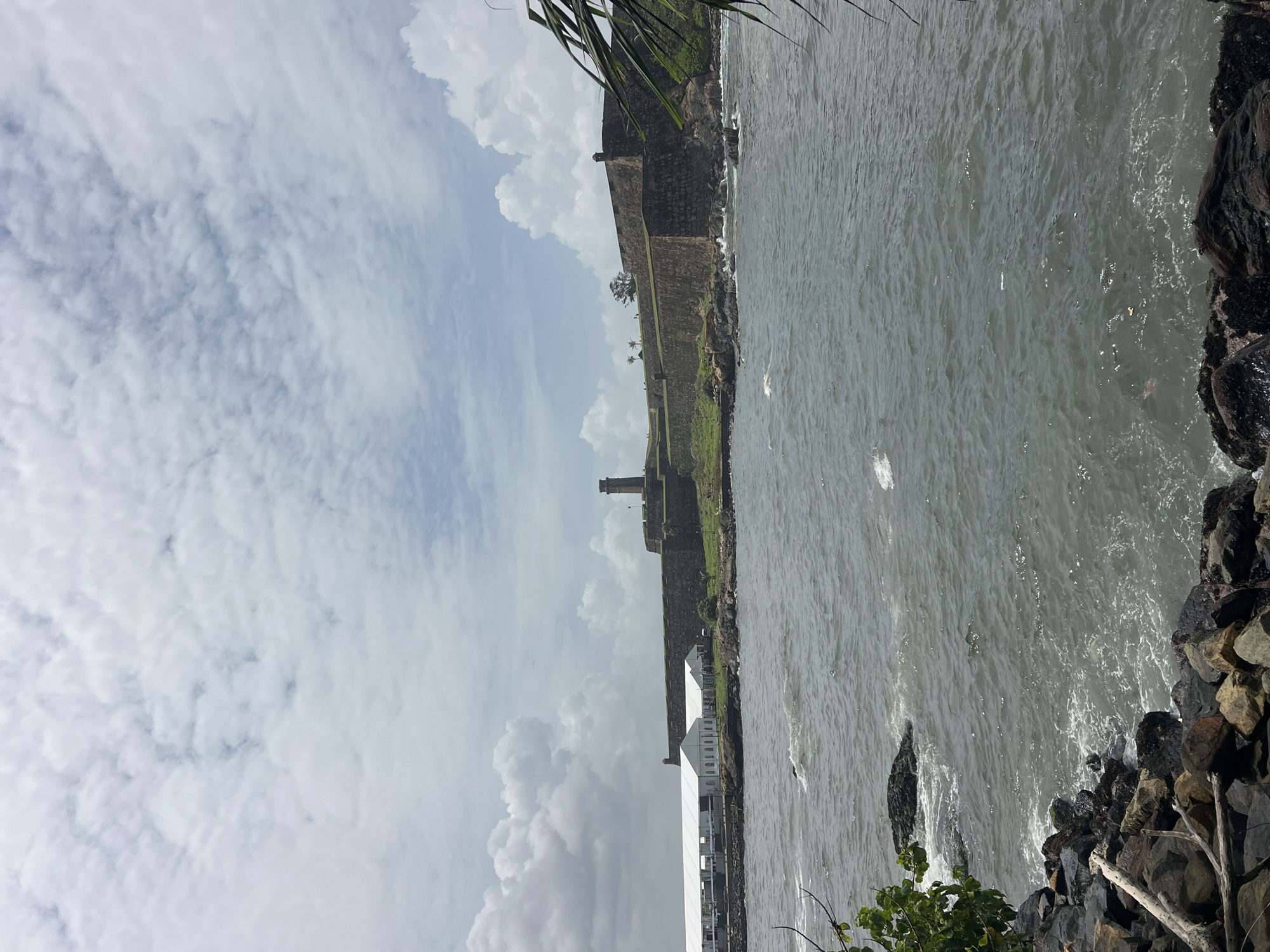 This photograph captures a view of Galle Fort, a UNESCO World Heritage site, from the ocean. The fort's imposing stone walls, topped with green grass and small watchtowers, stretch across the middle ground. A prominent lighthouse stands tall on the fort's ramparts. In the foreground, dark, rocky shorelines meet the choppy, grey ocean. White tents, likely temporary structures, are set up along the coast to the left of the fort. The sky is filled with a mix of bright white and grey clouds.