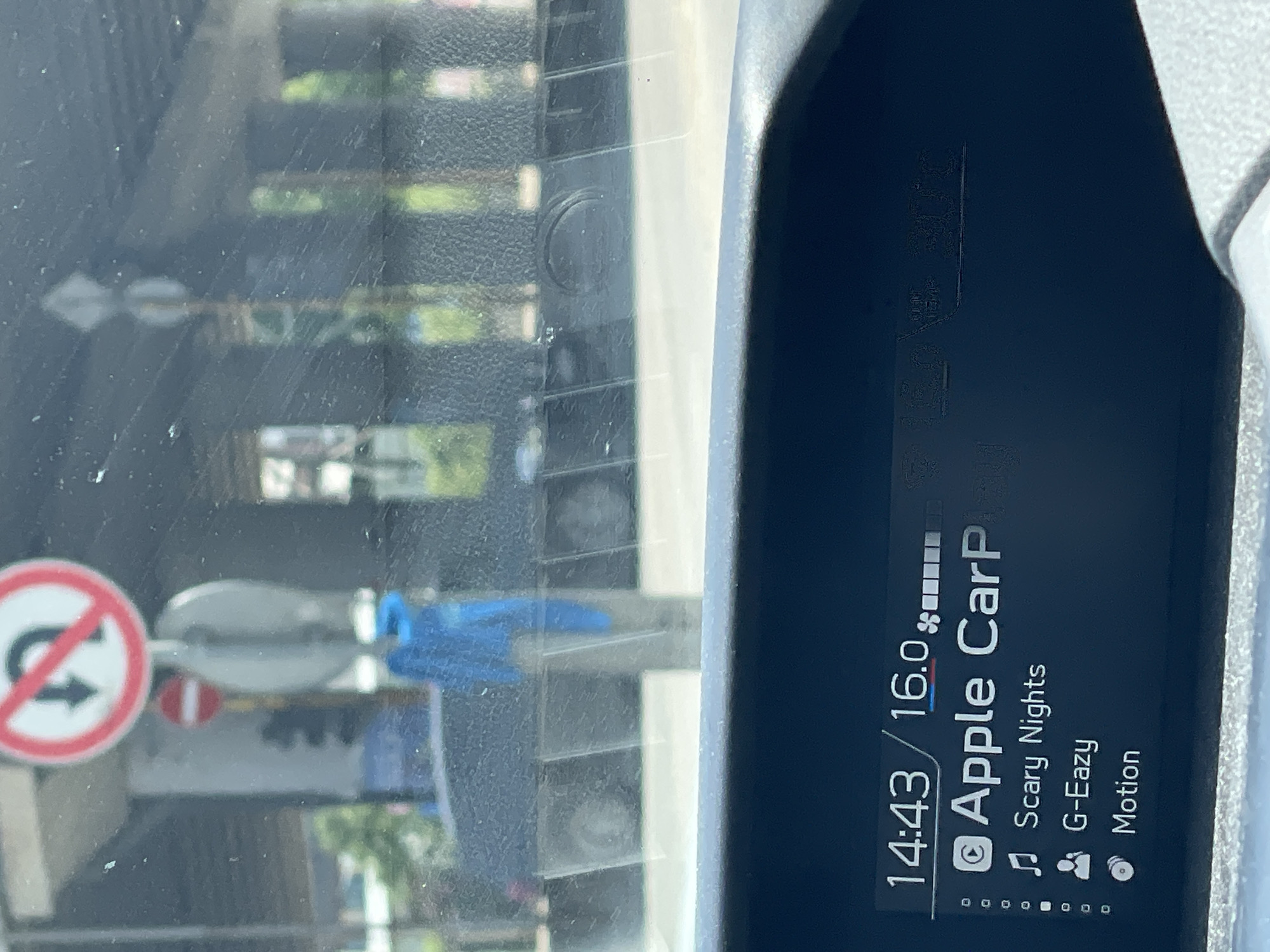 The image shows a car's dashboard display with the time "14:43" and temperature "16.0°C". The display indicates that "Apple CarPlay" is active, and the current song playing is "Scary Nights" by "G-Eazy", with "Motion" also visible. Outside the car, visible through the windshield, is a traffic sign prohibiting U-turns. The environment appears to be a street in Sofia, Bulgaria, with buildings and trees in the background. The windshield is dusty.