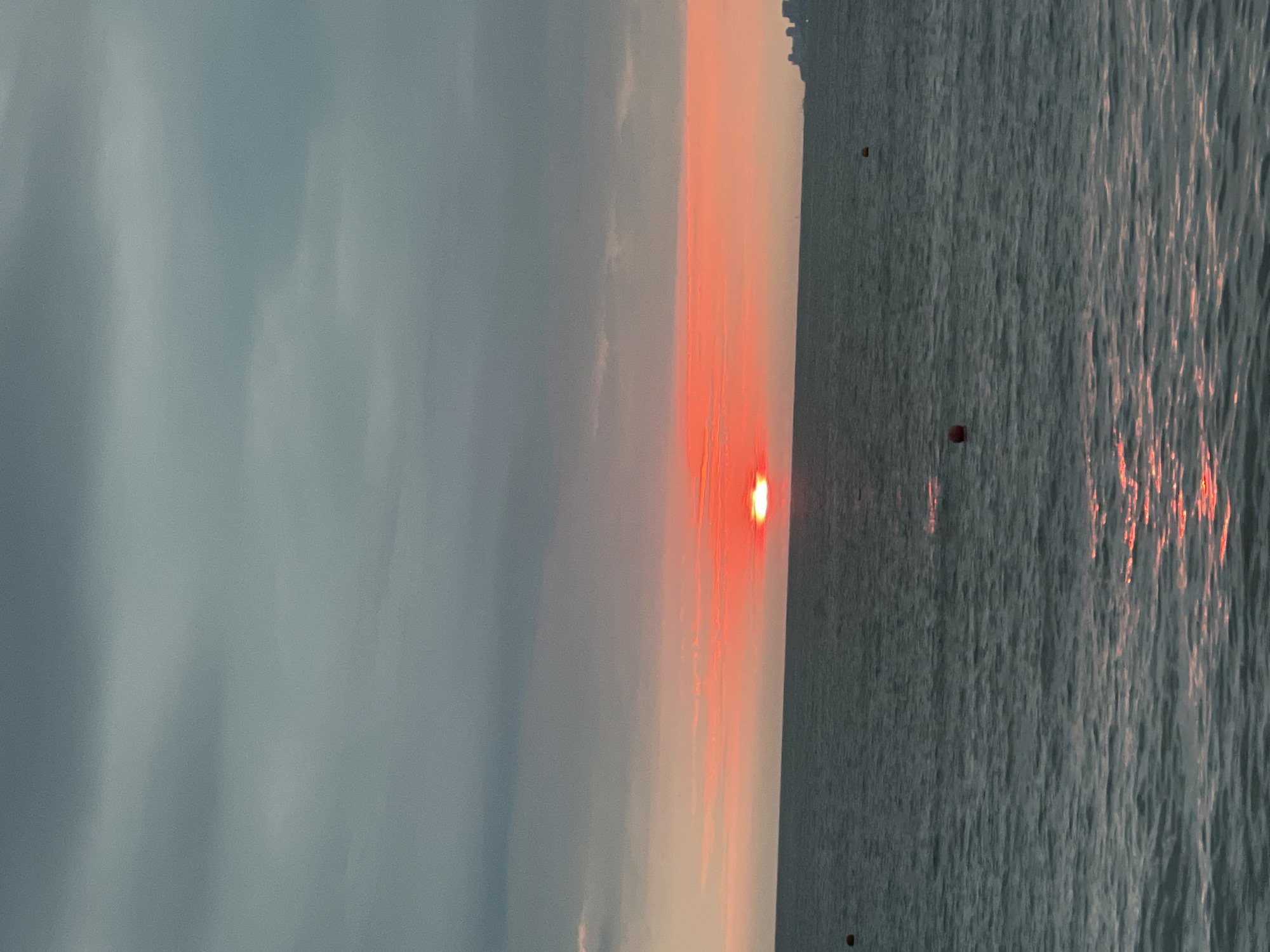 The image depicts a sunset over the Black Sea, with the sun appearing as a bright, red disc just above the horizon. The sky is filled with clouds, tinged with hues of orange and pink from the setting sun. The water is dark and choppy, with reflections of the sunset shimmering on its surface. In the distance, a city skyline is faintly visible on the right side of the frame, suggesting the location is near an urban area. Several buoys, one red and one orange, are scattered across the water. The overall atmosphere is serene and contemplative. This scene is likely from the city of Sochi, Russia, a popular Black Sea resort known for its beautiful coastal landscapes and sunsets.