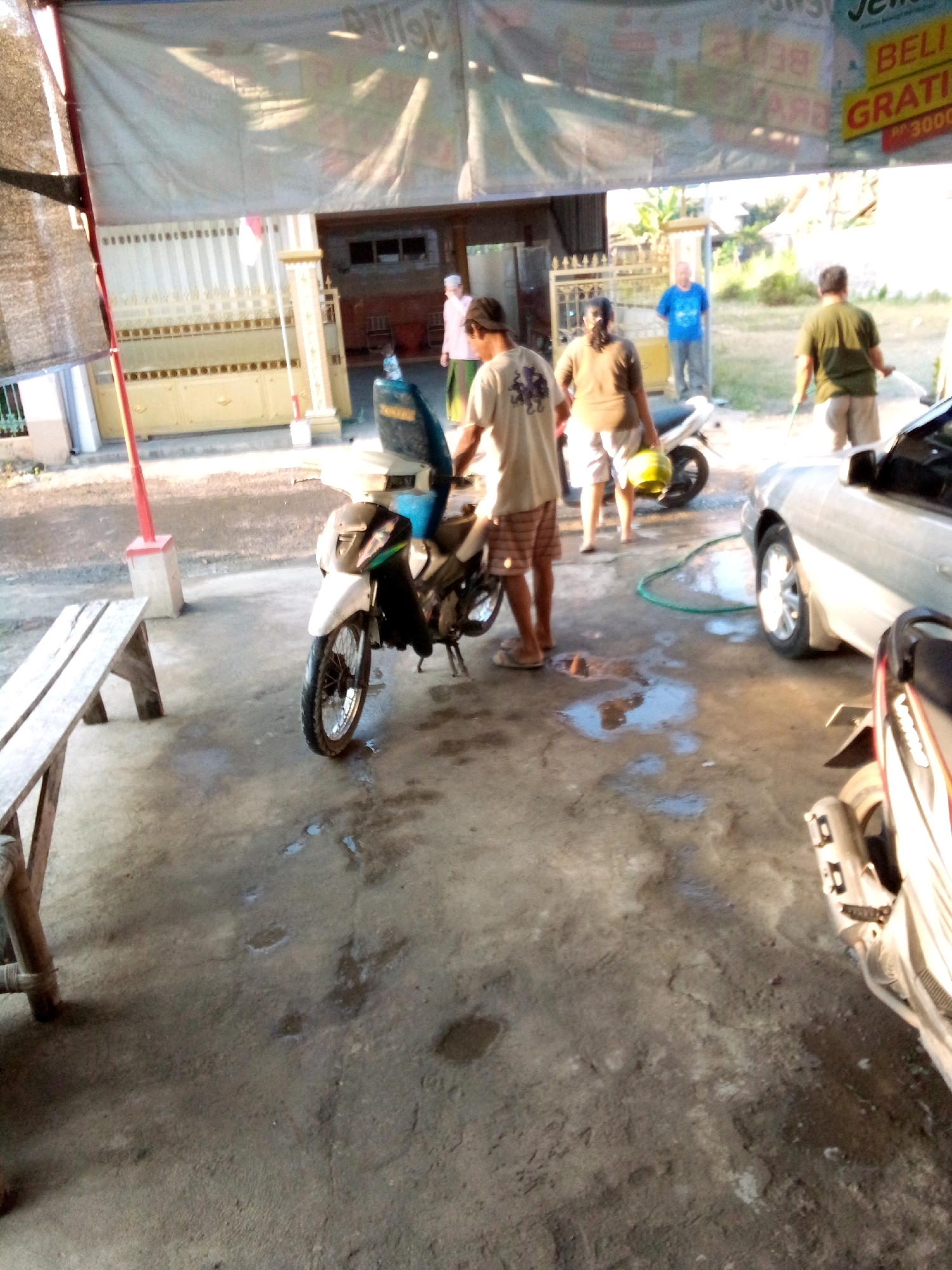 The image depicts an outdoor scene in Simo, Indonesia, likely a car wash or a similar service area. A white motorcycle is parked on a concrete surface, with its front wheel turned slightly to the left. The motorcycle appears to be clean, and there is some water on the ground around it, suggesting it might have been recently washed.
In the background, several people are visible. One person, a man wearing a light-colored t-shirt and plaid shorts, is standing next to the motorcycle, possibly attending to it. To the right of the motorcycle, a silver car is parked. A green hose is visible on the ground near the car, indicating active use of water.
Further in the background, there's a building with a closed gate and a banner hanging above it. The banner has text and graphics, with one section clearly displaying "GRATIS" and "RP 3000", suggesting a promotional offer. There are other individuals in the vicinity, including a person in a blue t-shirt standing near the gate and two people further back, one in a green shirt and another partially obscured. The overall environment appears to be sunny, with shadows cast on the ground. A wooden bench is positioned to the left of the motorcycle.