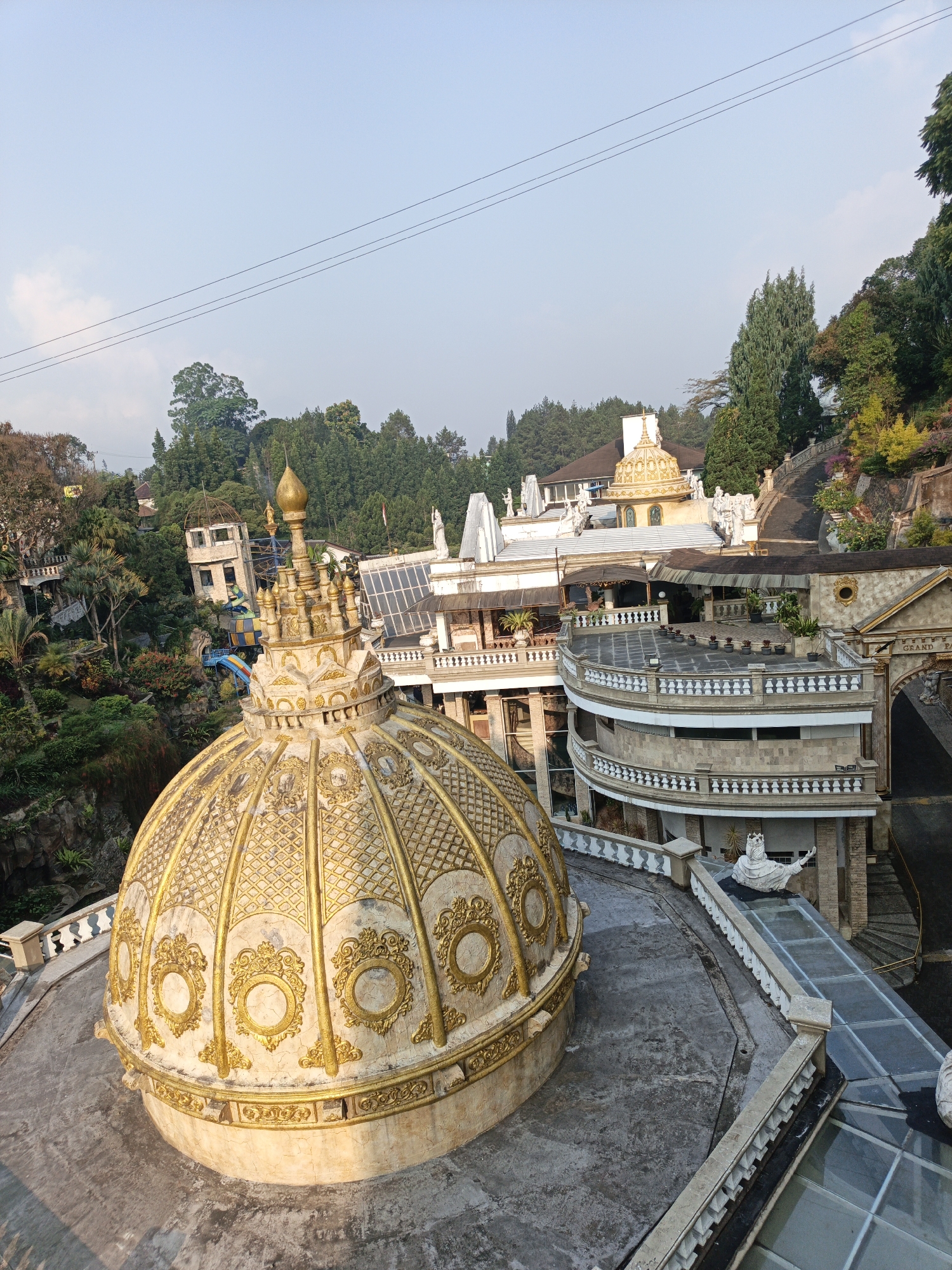 The image captures a detailed view of a multi-tiered, ornately decorated building complex, likely a resort or tourist attraction, nestled within a lush, green, hilly landscape. In the foreground, a large, golden dome dominates the scene, featuring intricate patterns, decorative circular motifs, and a pointed finial. The dome rests on a concrete platform.
Behind the dome, the architecture unfolds in a series of balconies, terraces, and distinct building sections. Many of these structures are adorned with white balustrades and classical-inspired statues, adding to a grand, somewhat whimsical aesthetic. One notable building features a smaller golden dome. The complex appears to be built into the side of a hill, with pathways and stairs winding through the property.
Surrounding the buildings is a dense forest, with a variety of trees and vegetation, some of which appear to be landscaped gardens with colorful flowers. A water slide is visible amongst the greenery on the left side of the image. Power lines stretch across the pale blue sky, indicating the presence of infrastructure.
The overall impression is of a meticulously designed, themed environment, likely a destination for leisure and entertainment, situated in a natural, elevated setting. The location is identified as Cibogo, Indonesia. The image is taken from a high vantage point, looking down and across the complex, suggesting the photographer is on an upper level of the same or an adjacent structure. There are no people visible in the image.