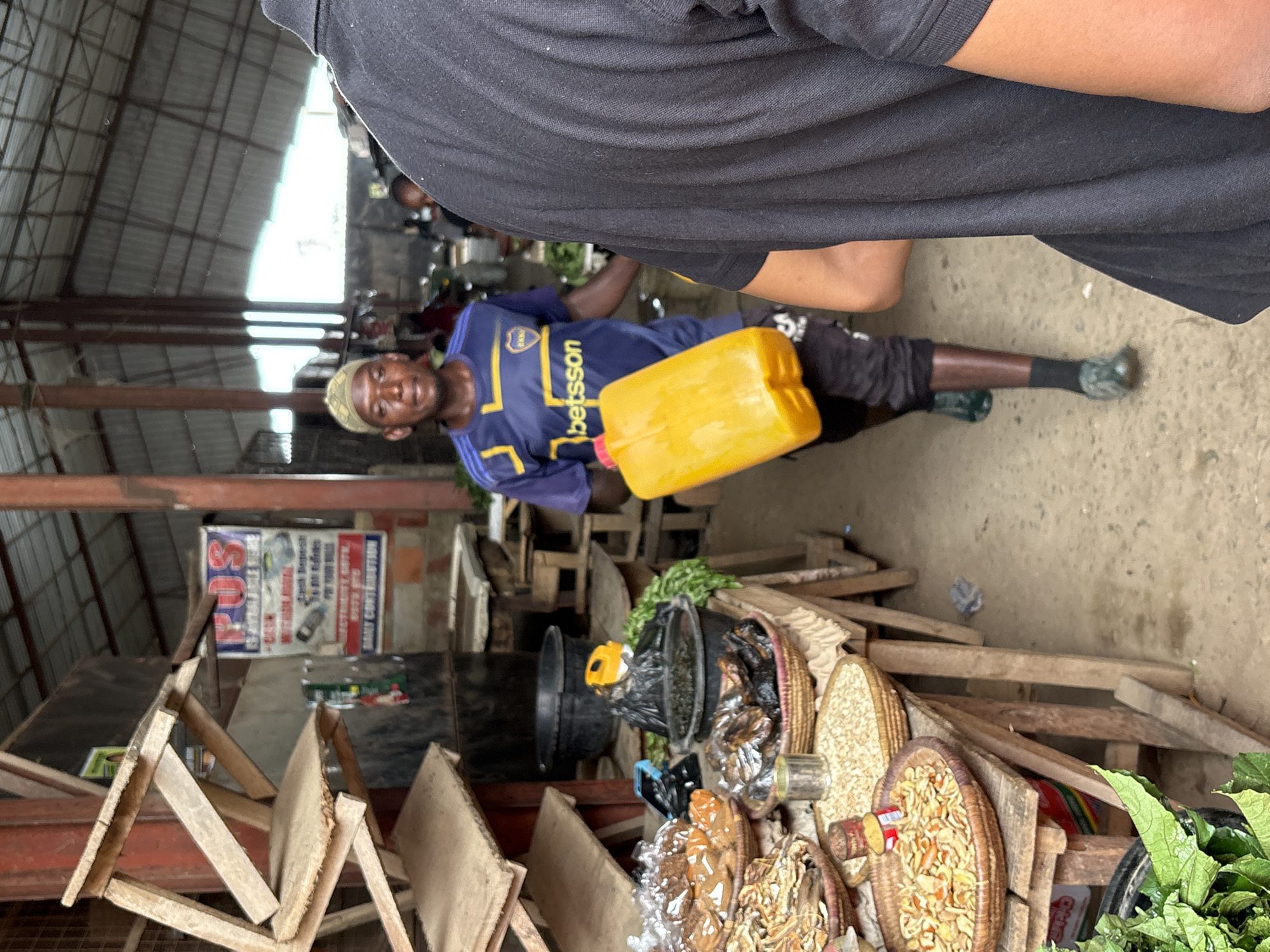 The image depicts a scene inside what appears to be a market or a commercial area. A man is holding a yellow plastic container. He is wearing a blue and yellow Betsson branded shirt. He is also wearing shorts, and socks with green boots. Other objects include stacked wooden crates, various baskets containing food items, a black plastic bucket, and fresh green leafy vegetables. A blurry sign hangs in the background. The setting is Iddo Tudunwada, Nigeria.