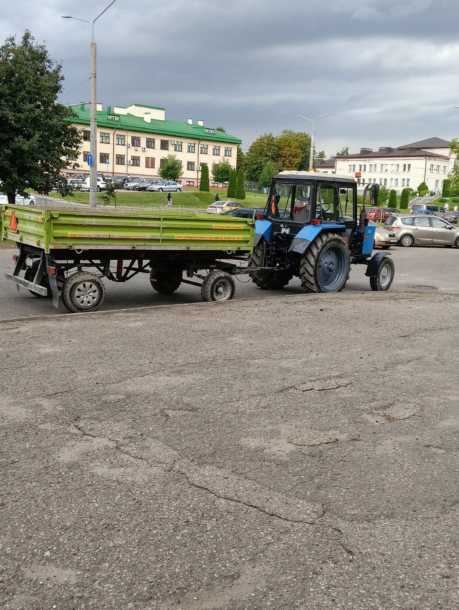 In Hrodna, Belarus, on a cloudy day, a blue tractor is parked next to a green trailer on a cracked asphalt surface. The tractor has a black cab and a license plate that reads "9842 HA-5". The trailer is long and rectangular with two sets of dual wheels on each side. The trailer has yellow warning stickers and some text in Cyrillic. In the background, there are several buildings with green roofs, trees, and parked cars. A street light stands tall to the left of the tractor.