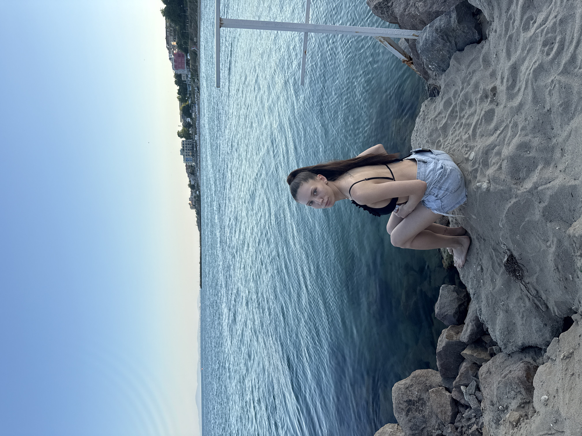 A young woman is crouched on a rocky shoreline with a sandy patch in front of her. She is wearing a black bikini top and light denim shorts. Her long, dark hair is pulled back in a ponytail. Her gaze is directed at the camera with a neutral expression. The water is calm and blue, with a faint haze in the distance where a shoreline with buildings is visible. The sky is a clear, pale blue, indicating it is likely daytime, possibly late afternoon or early evening due to the soft light. Large, dark rocks frame the foreground and extend into the water. A white metal structure is partially visible in the upper left portion of the frame, suggesting a pier or a similar waterfront installation. The overall scene is serene and suggests a moment of rest or posing by the water.