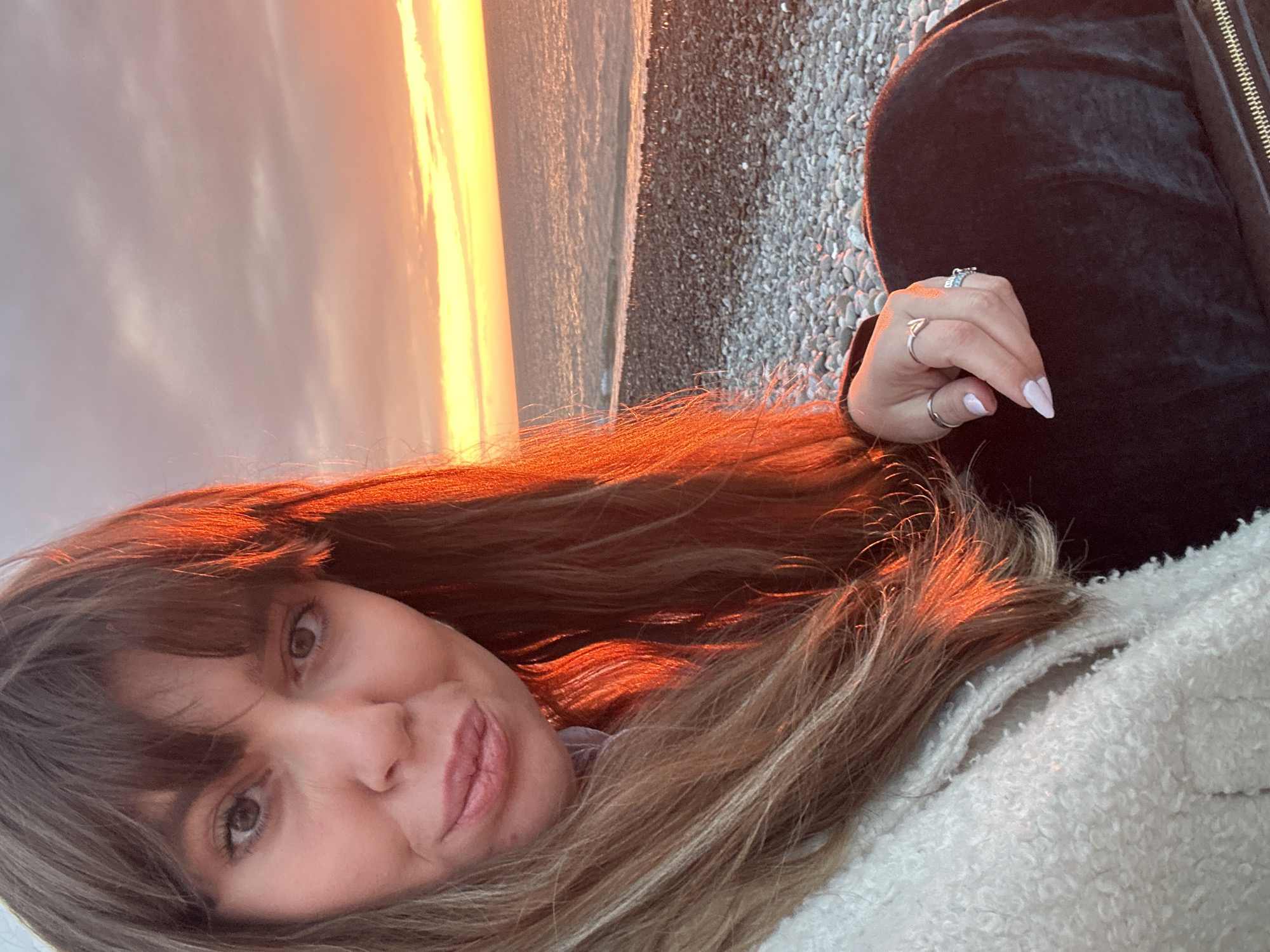 A selfie taken on the coast of Batumi, Georgia, at sunset. A woman with brown hair and light skin is visible. Her hair is partially illuminated by the orange light of the setting sun. She is wearing a white, textured jacket and a black velvet jacket. Several rings are visible on her fingers. In the background, the ocean meets the shore. The sky is illuminated by a bright orange sunset. Gray pebbles are visible on the beach.