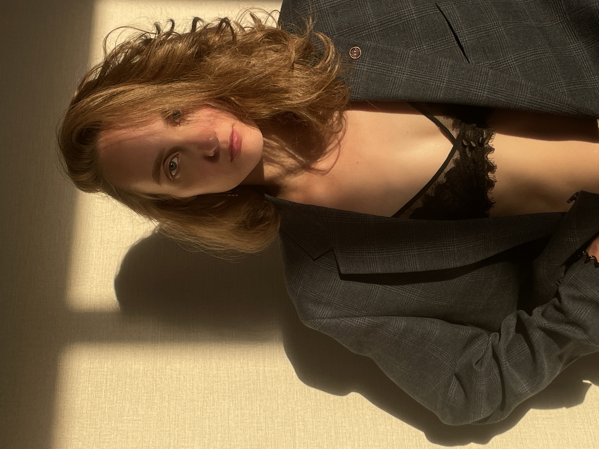 A fair-skinned woman with auburn hair lies on a surface with a textured, off-white covering. She wears a dark gray plaid blazer partially open, revealing a black lace bra. Sunlight creates strong shadows on the surface, including a shadow of the woman's head and torso. The location cannot be confirmed, as no contextual information points to Подшивалово, Russia.